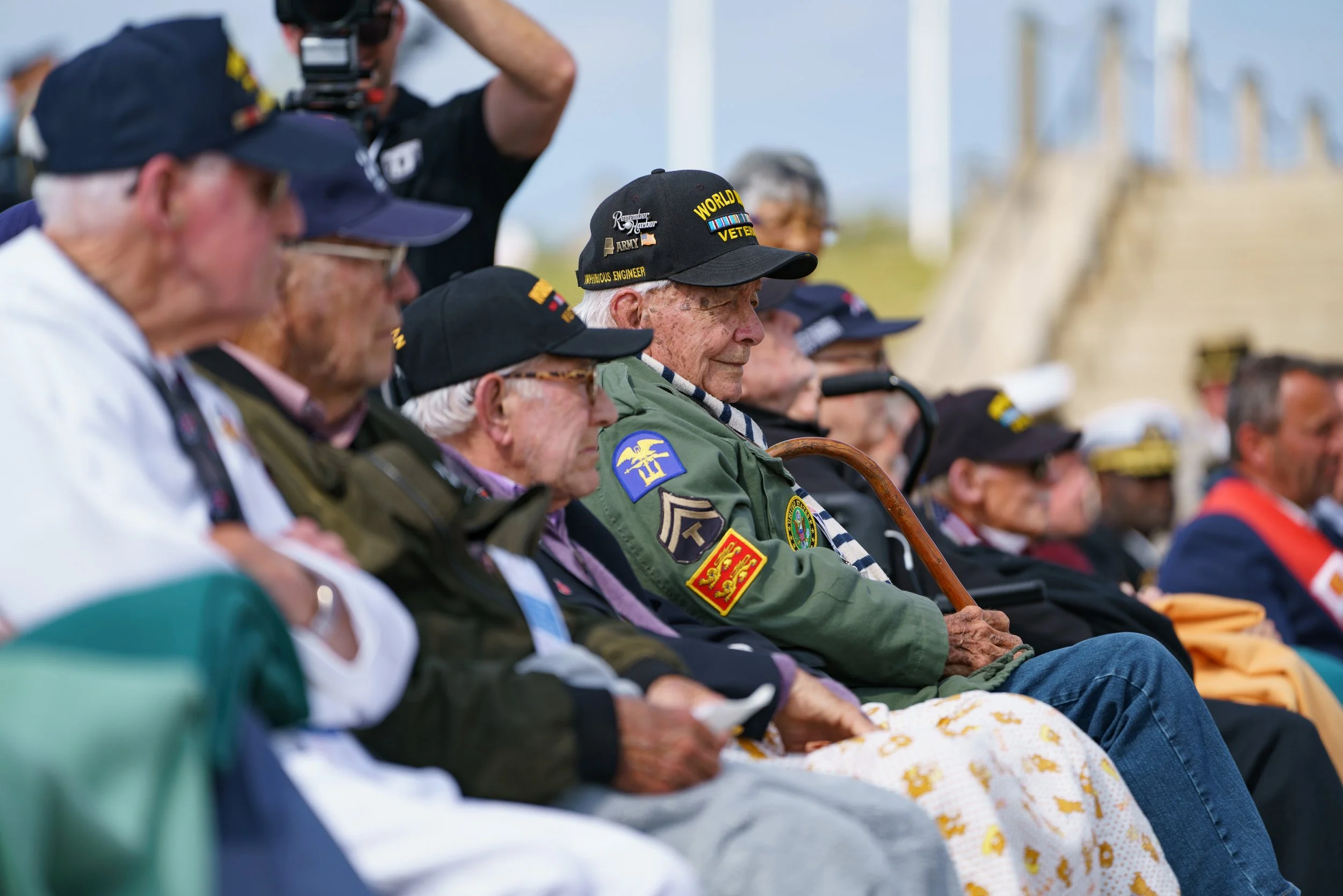 Veterans sitting in a row outdoors, wearing military hats with patches and insignia, some with canes, attending a remembrance event.