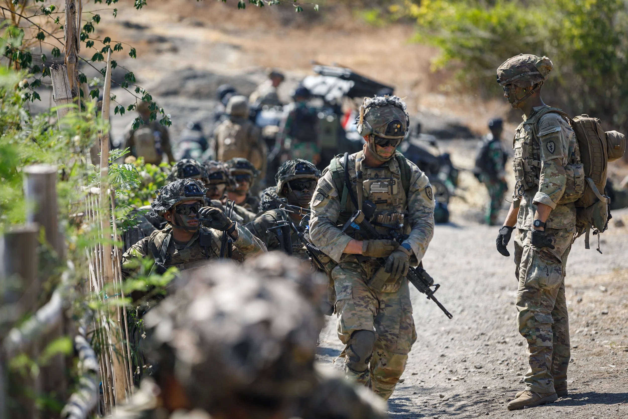 Military soldiers in camouflage uniforms gathered outdoors on a dusty path, some kneeling behind a makeshift barrier, while others are standing and talking, with military vehicles in the background.
