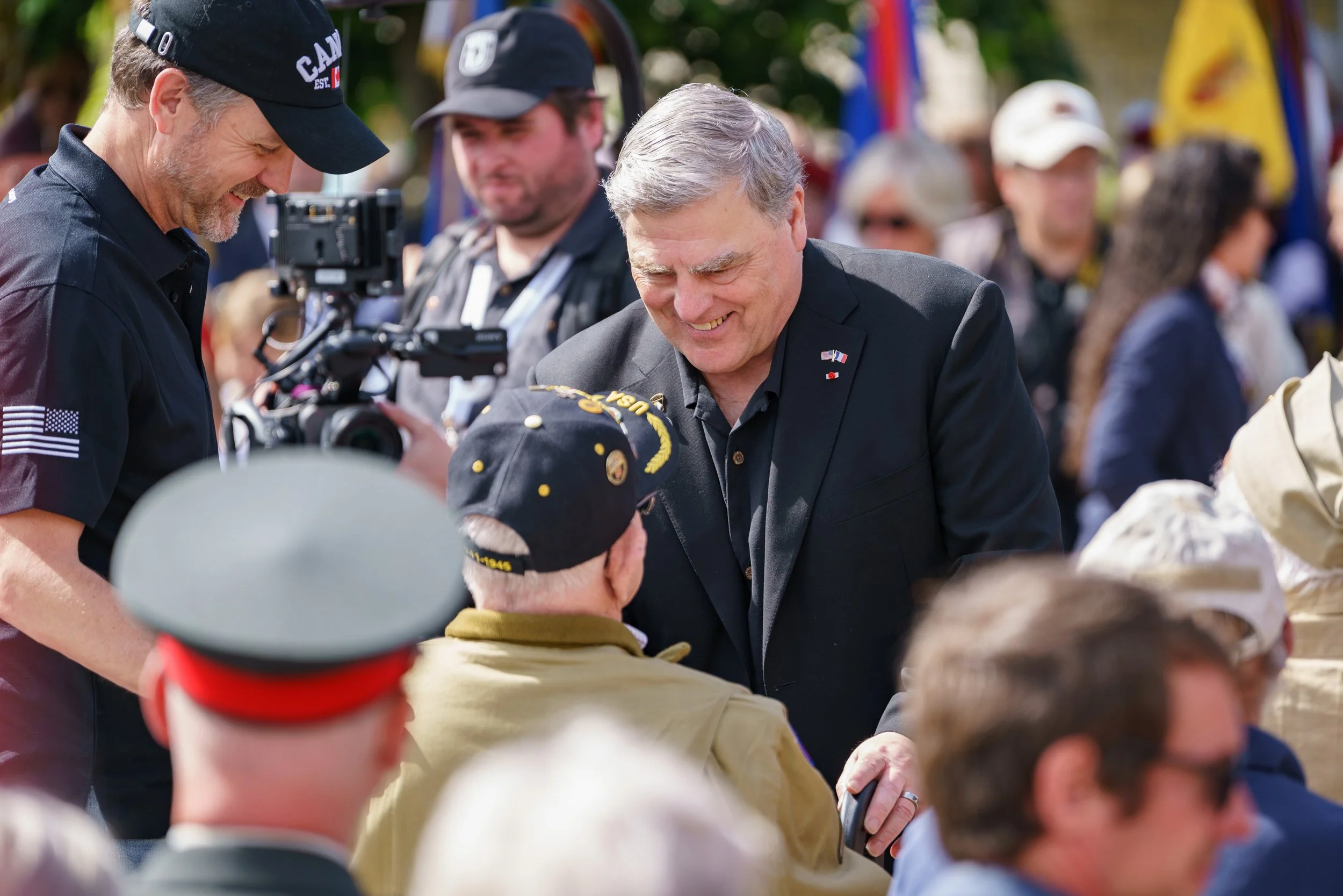A man in a black suit and gray hair smiling and talking to a veteran wearing a black veteran hat. They are at an outdoor event with many people in the background.