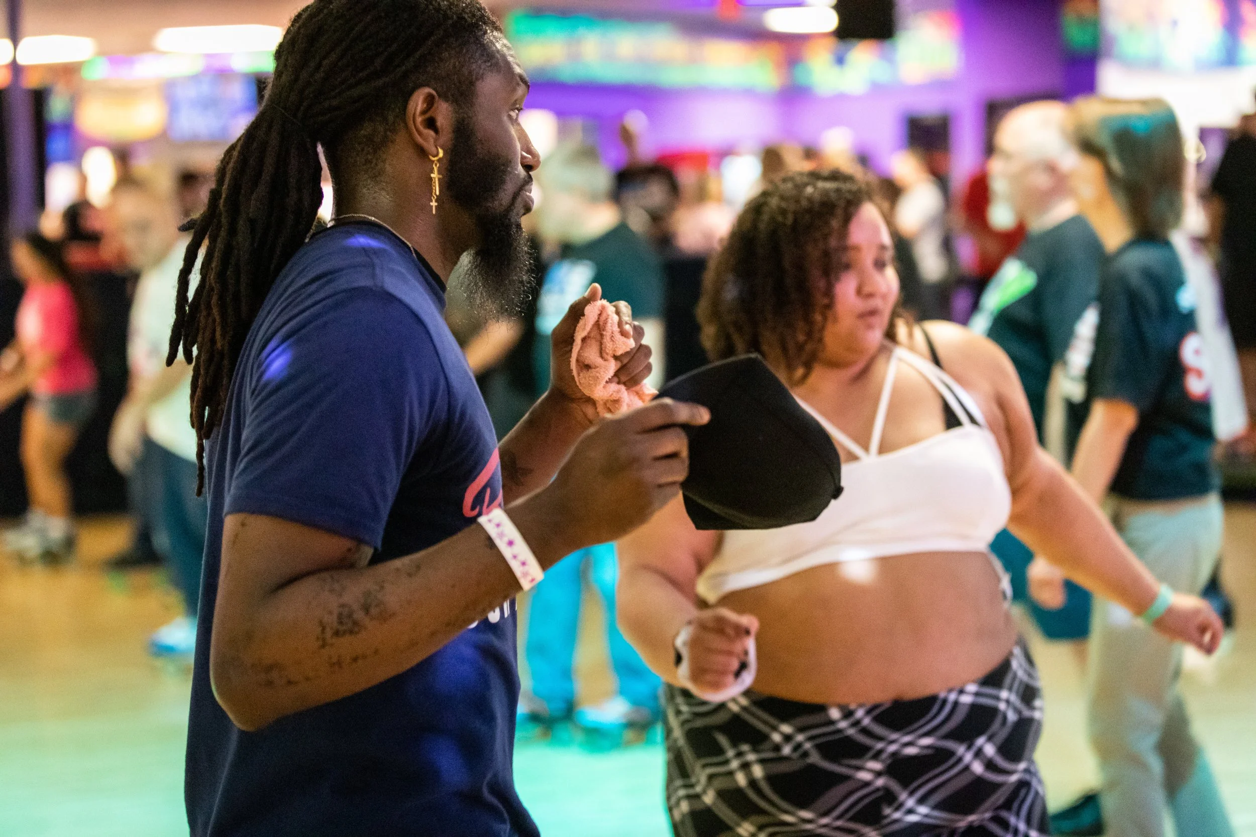 A man with long dreadlocks and earrings and a woman in a crop top dance at an indoor event, with colorful lights and other people in the background.