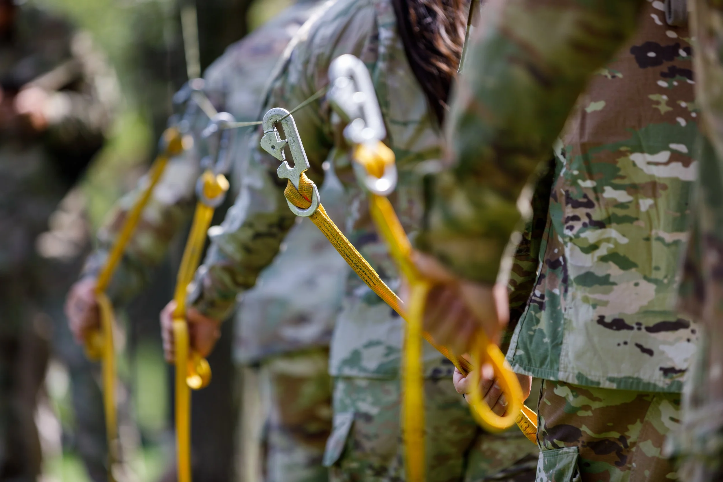 Close-up of soldiers in camouflage uniforms synchronized in a training exercise, holding a yellow safety harness and carabiners linked together.