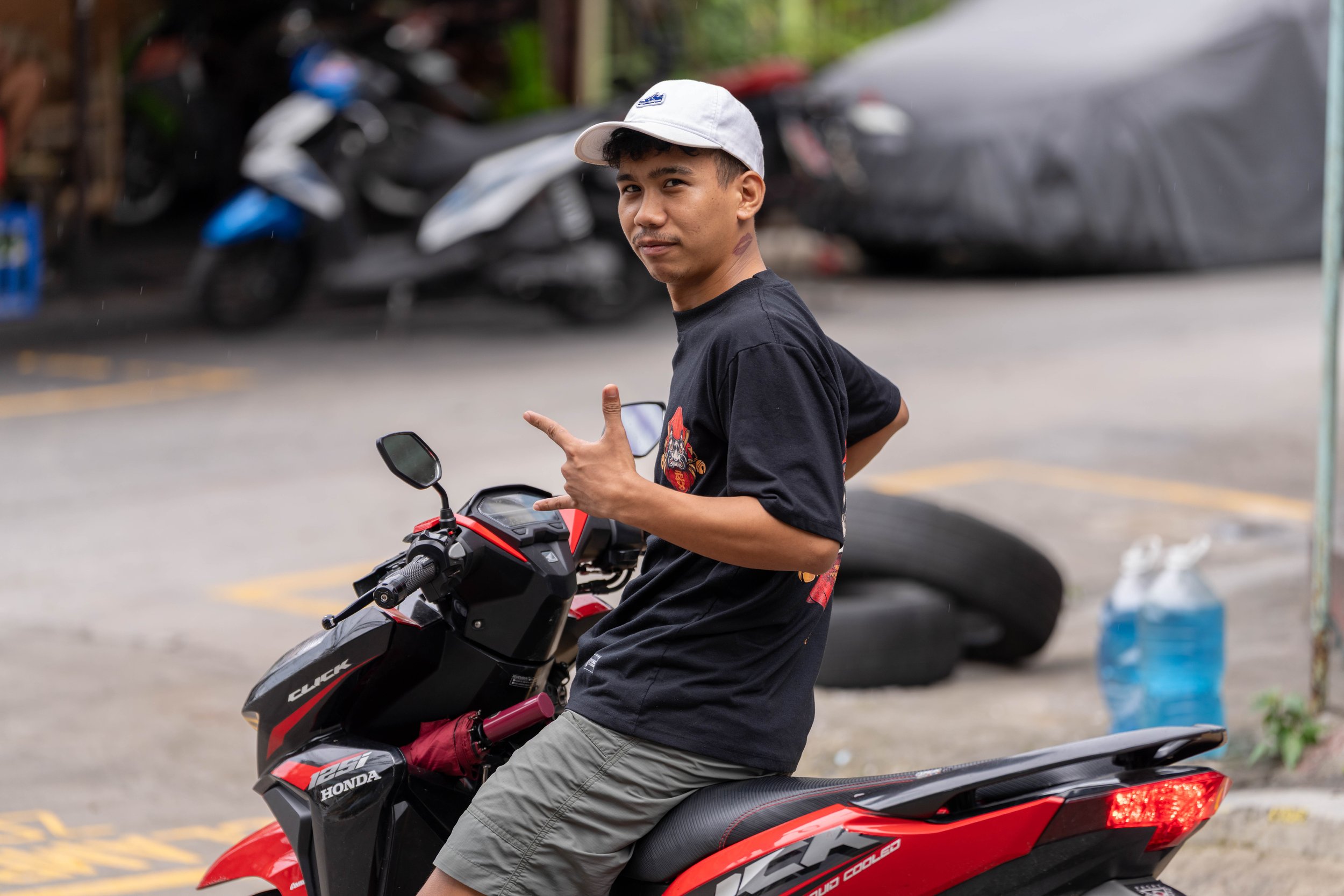 A young man in a black T-shirt and gray shorts sitting on a red and black Honda scooter, making a peace sign with his right hand while looking at the camera, wearing a white cap, with motorcycles and tires in the background.