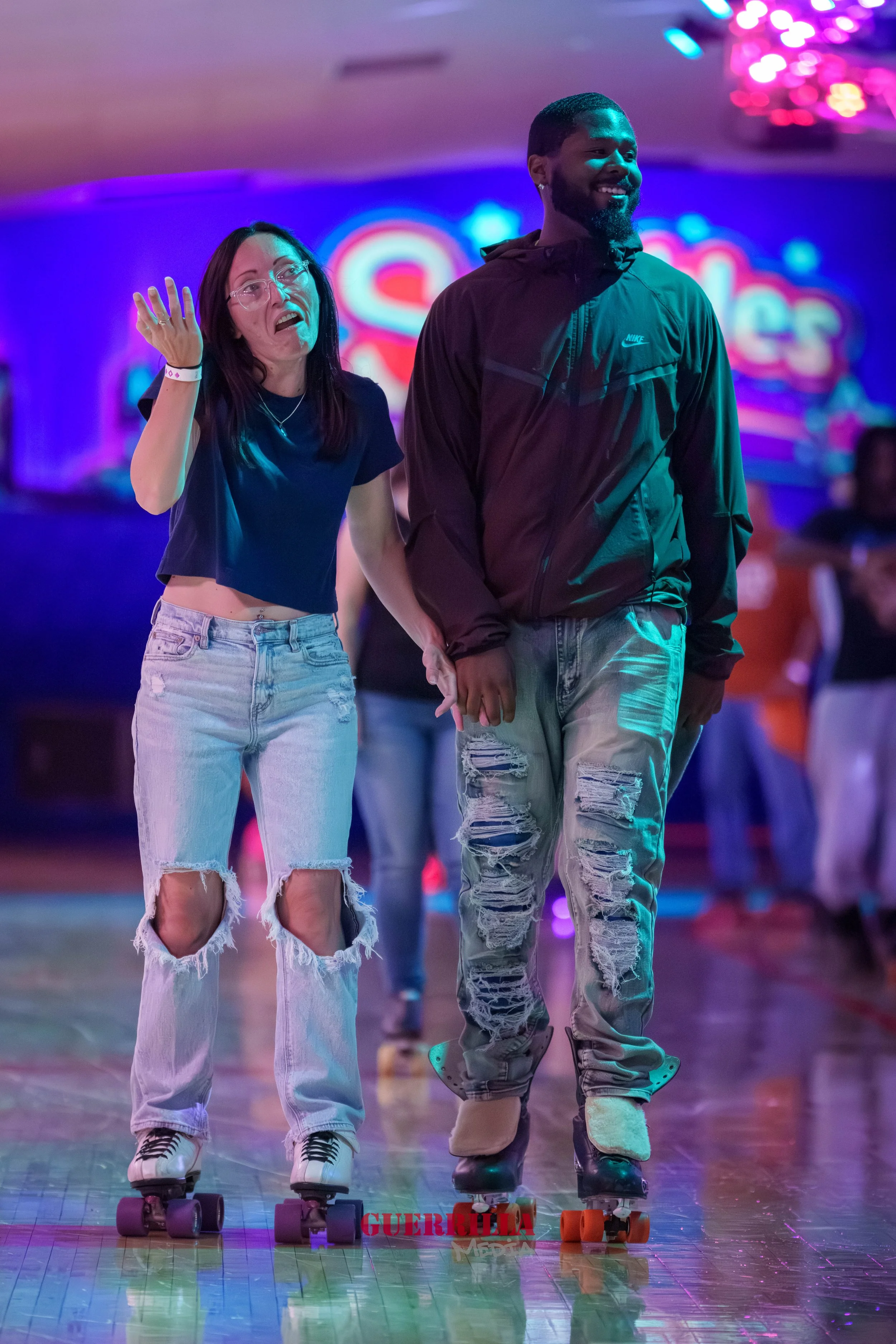 A couple roller skating together in a brightly lit indoor skating rink, holding hands and smiling, with colorful neon lights in the background.