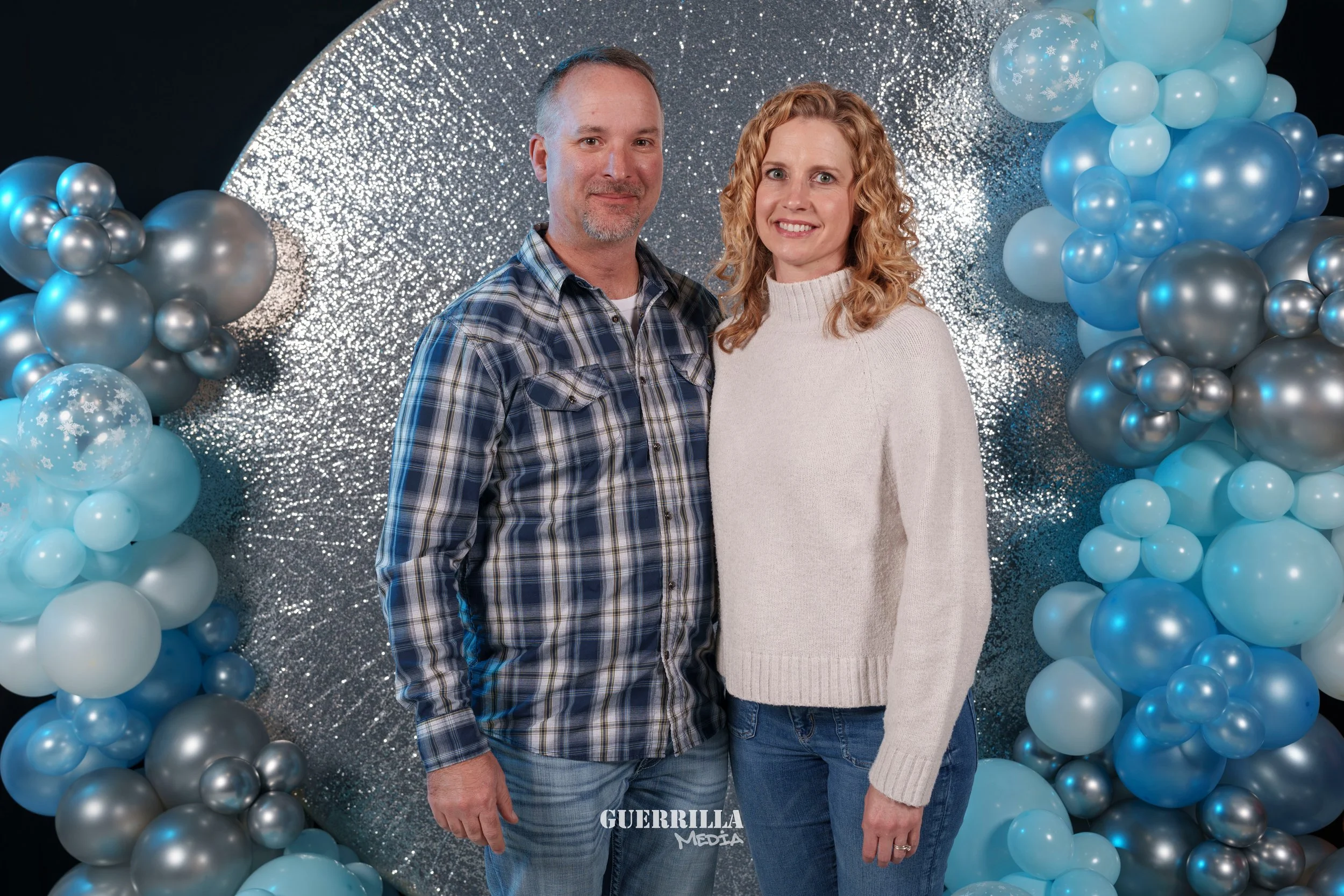 A man and woman standing together in front of a glittery and balloon backdrop. The man is wearing a plaid shirt, and the woman is wearing a white sweater. They are smiling and facing the camera.