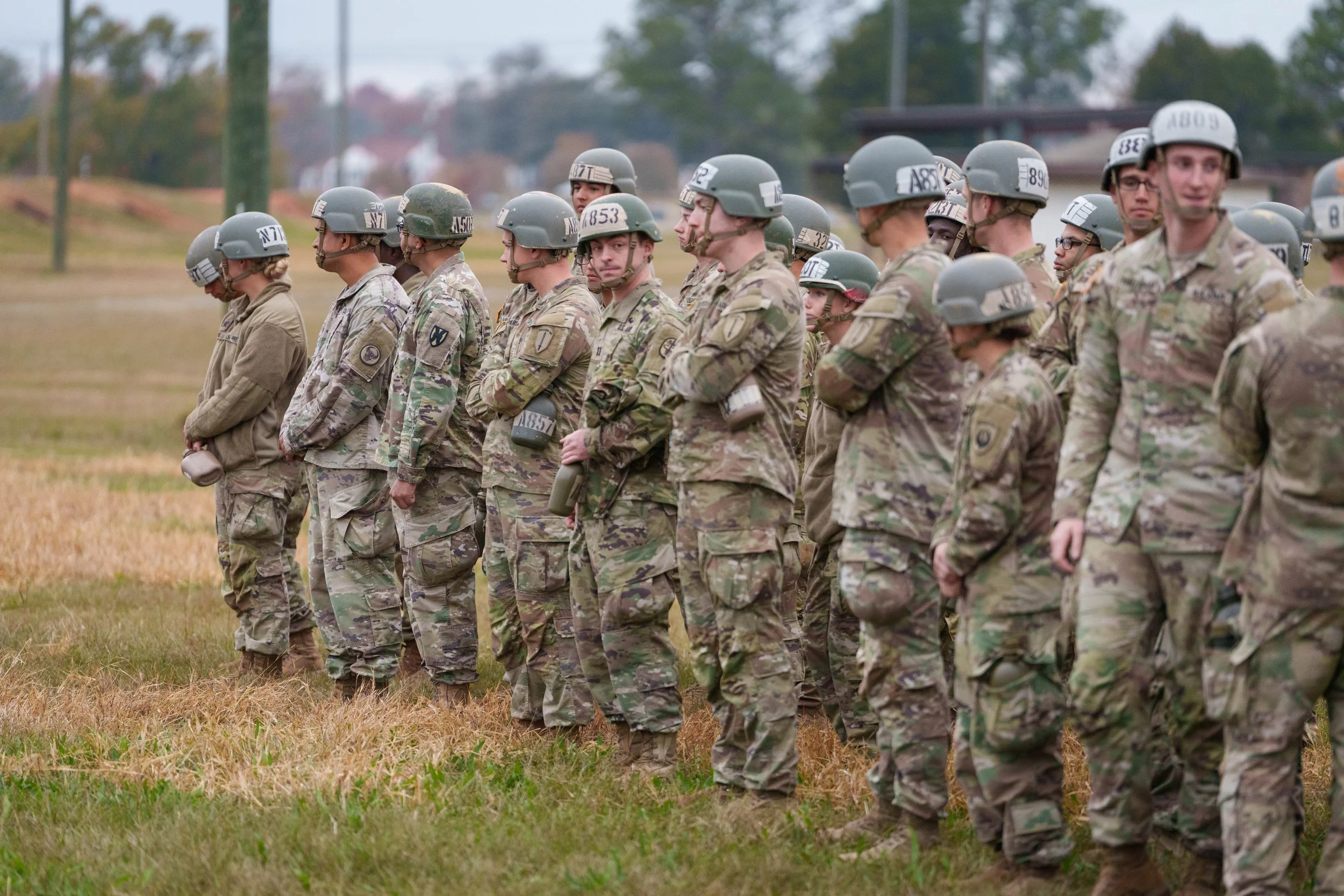 Group of soldiers standing in formation outdoors on a grassy field, wearing camouflage uniforms and helmets.