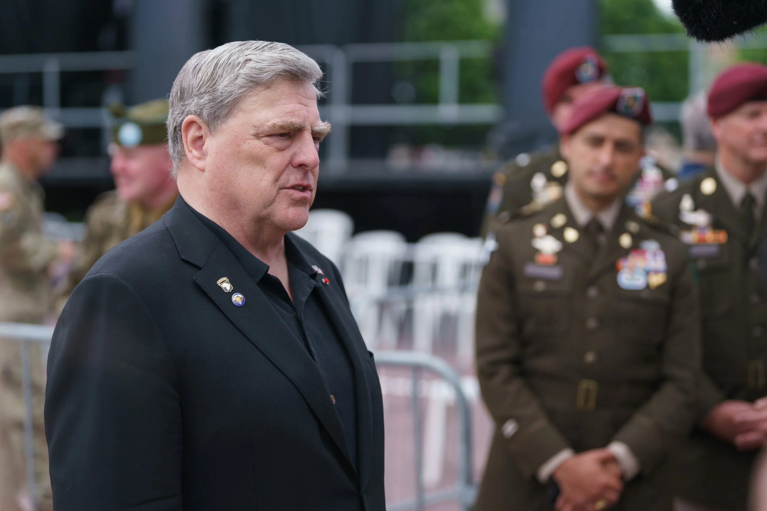 A man in a black suit with military pins on his lapel stands solemnly in front of a group of soldiers in uniform. The soldiers are wearing medals and maroon berets, with others blurred in the background.