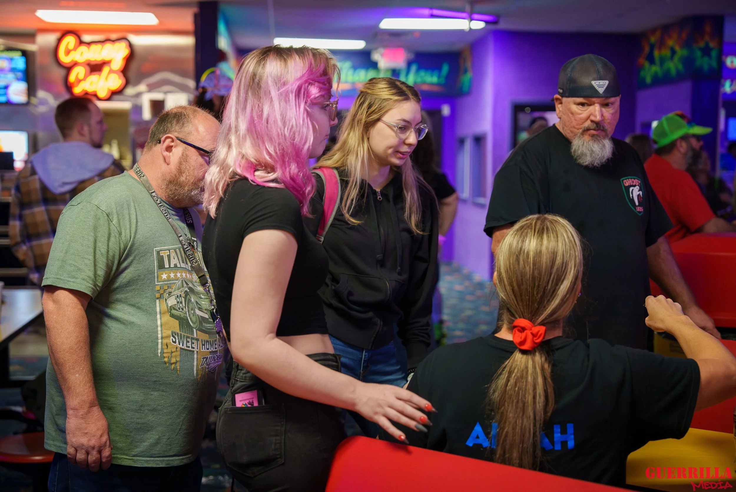 A group of five people, three women and two men, gathered around a seated woman with blonde hair tied back with a red scrunchie in an arcade or entertainment center. The woman standing with pink-tinted hair is touching the seated woman's arm, and the