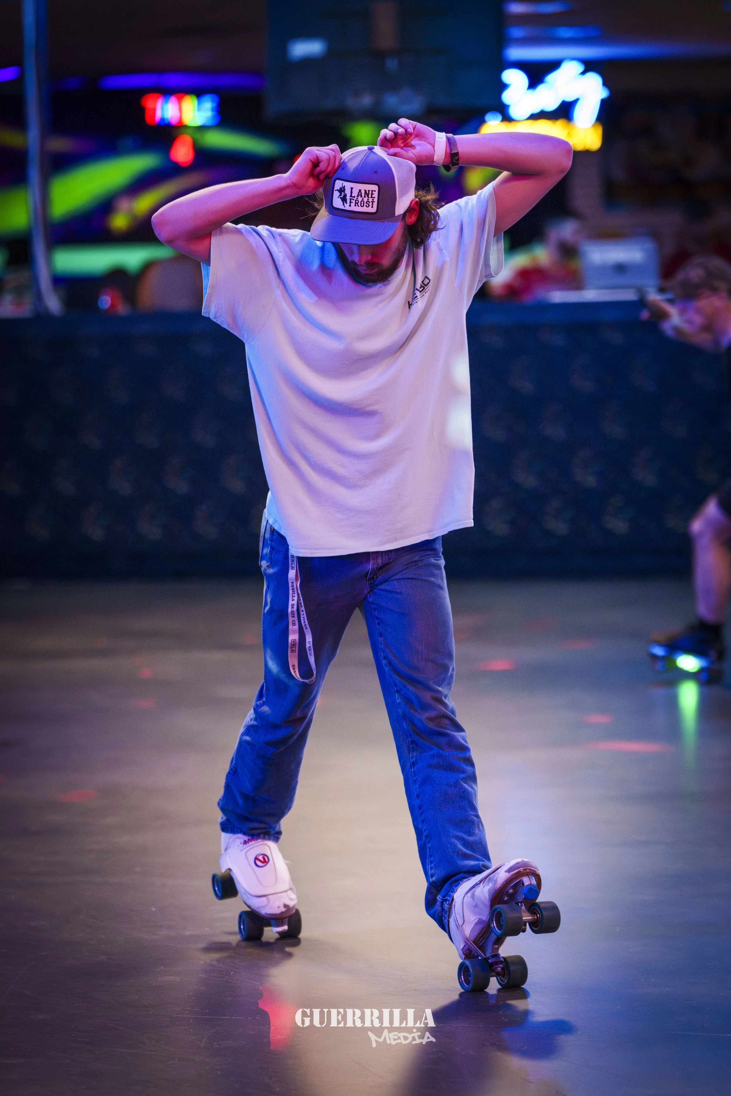 A man roller skating in a neon-lit arcade or skating rink, wearing a white t-shirt, jeans, a cap, and protective wristbands.