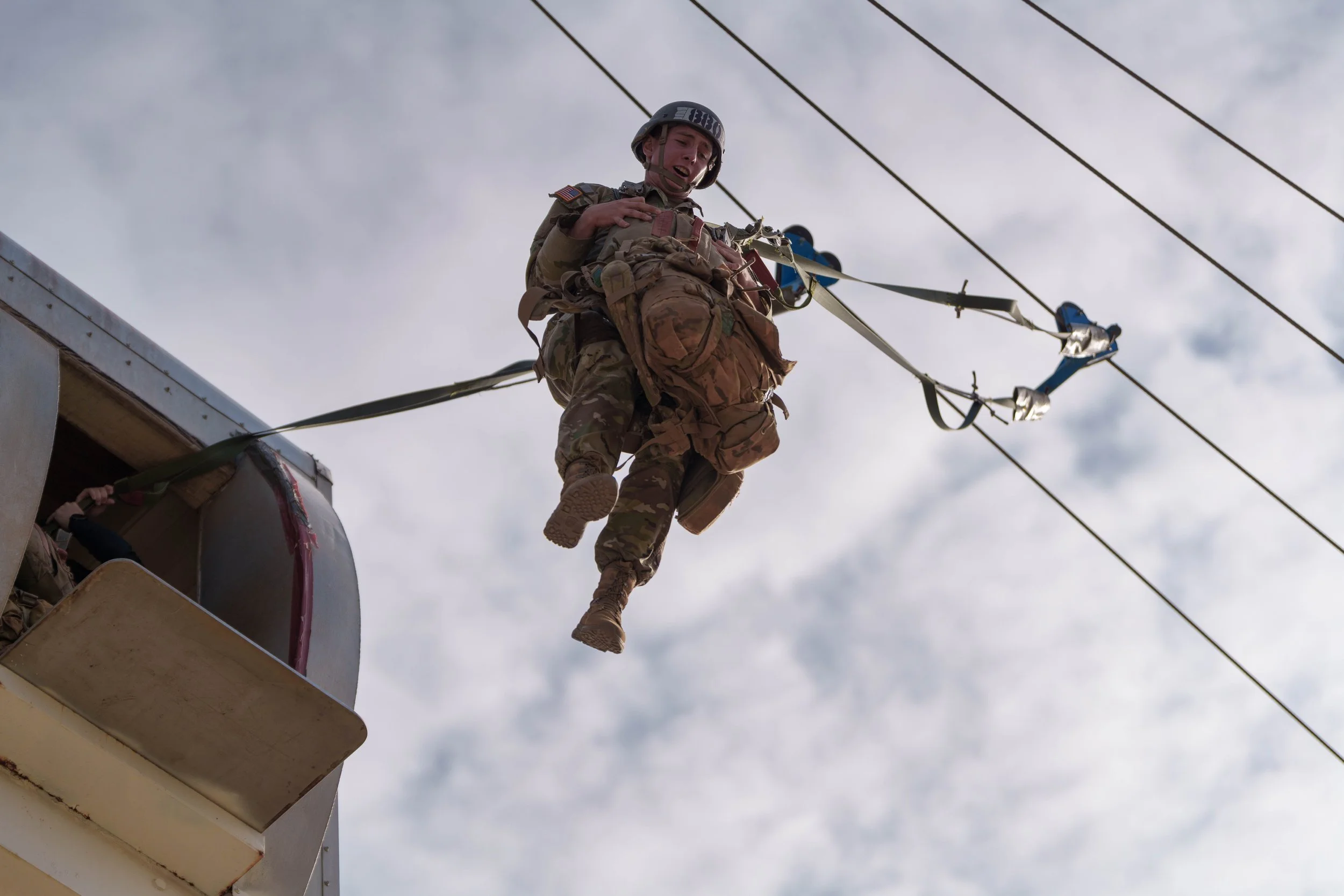 A soldier dressed in camouflage gear and helmet is being lowered from a military helicopter by a harness attached to cables, with a cloudy sky in the background.
