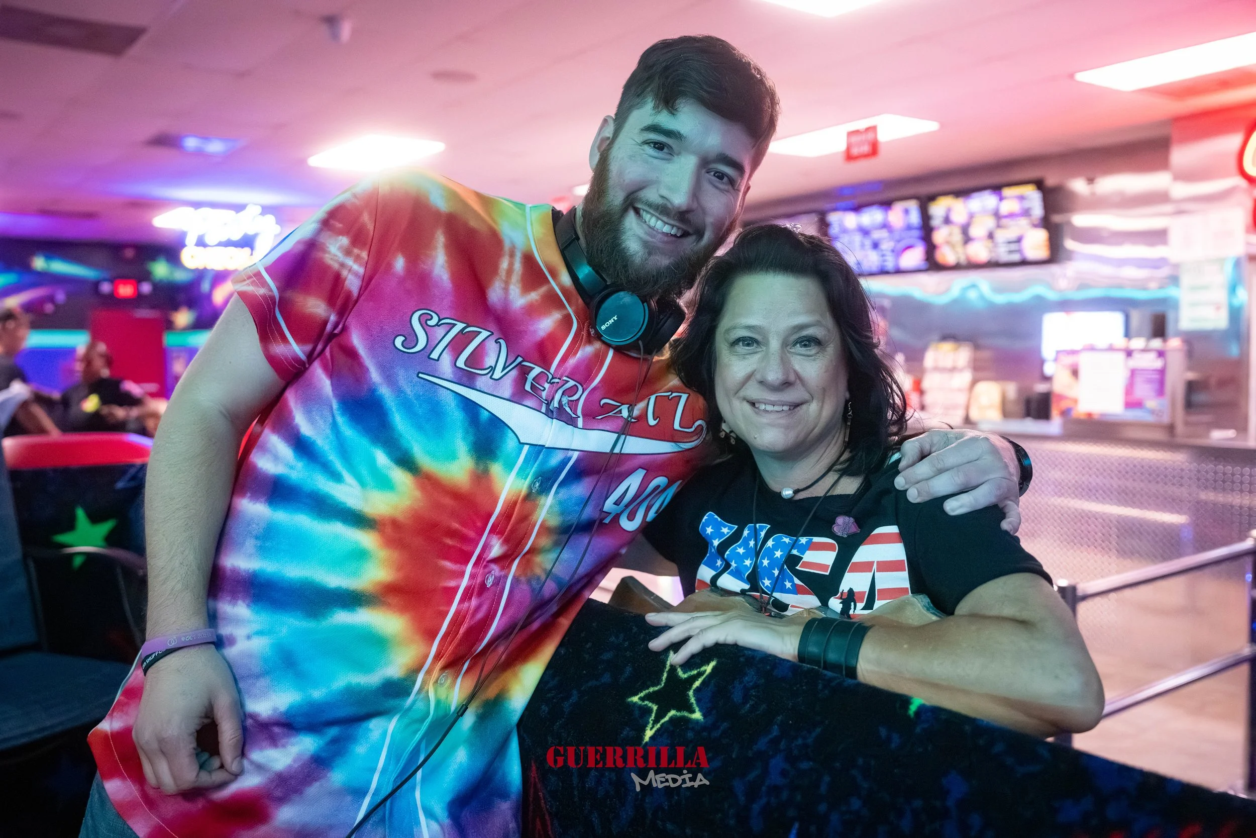 A young man with a beard and a woman with dark hair standing together at a bowling alley, smiling at the camera. The man is wearing a colorful tie-dye shirt and headphones, and the woman is wearing a black t-shirt with a patriotic design. The backgro