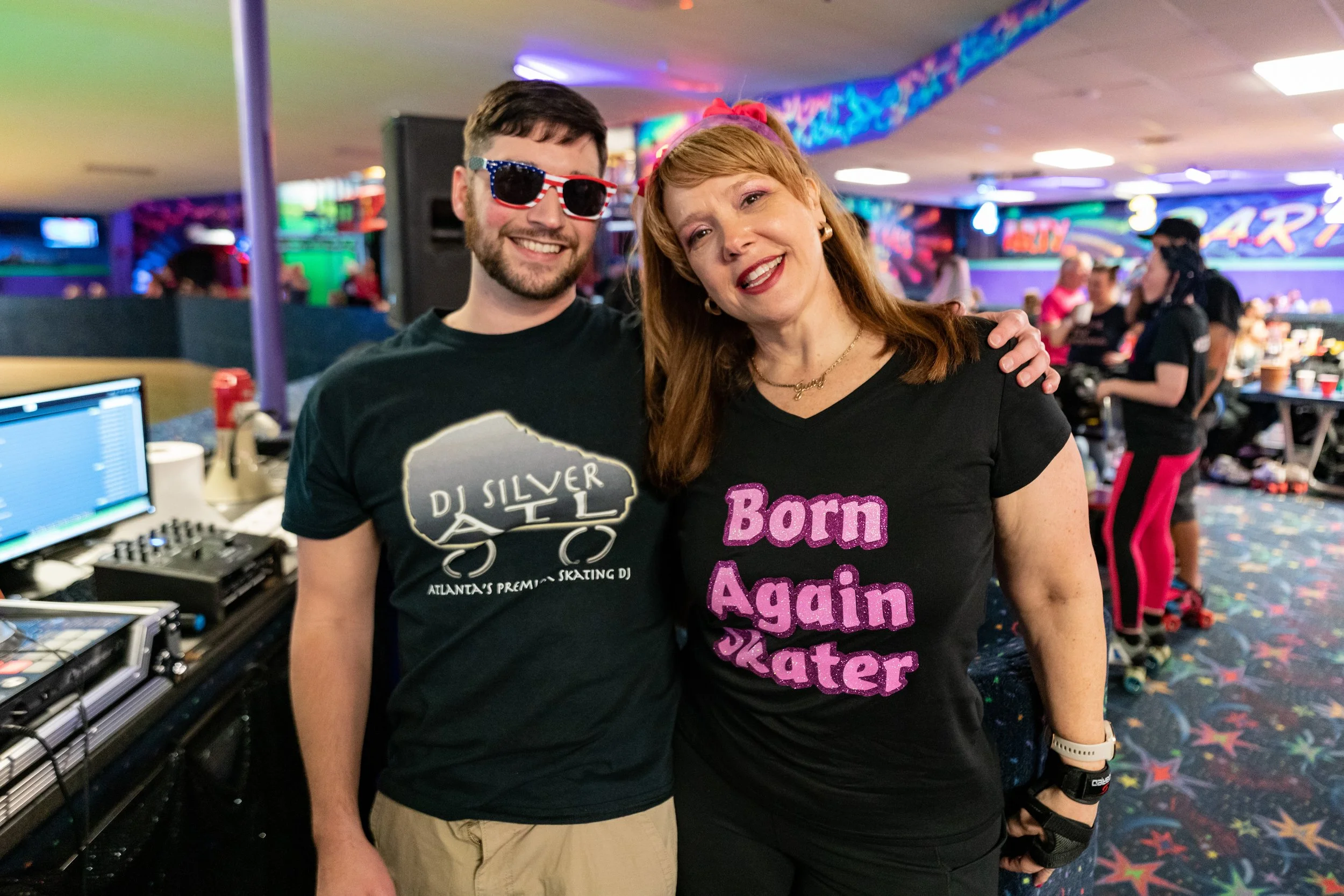 A man and a woman posing together at a roller skating rink. The man is wearing sunglasses with an American flag design and a black t-shirt with DJ Silver's logo. The woman is wearing a black t-shirt that says 'Born Again Skater' in pink letters.