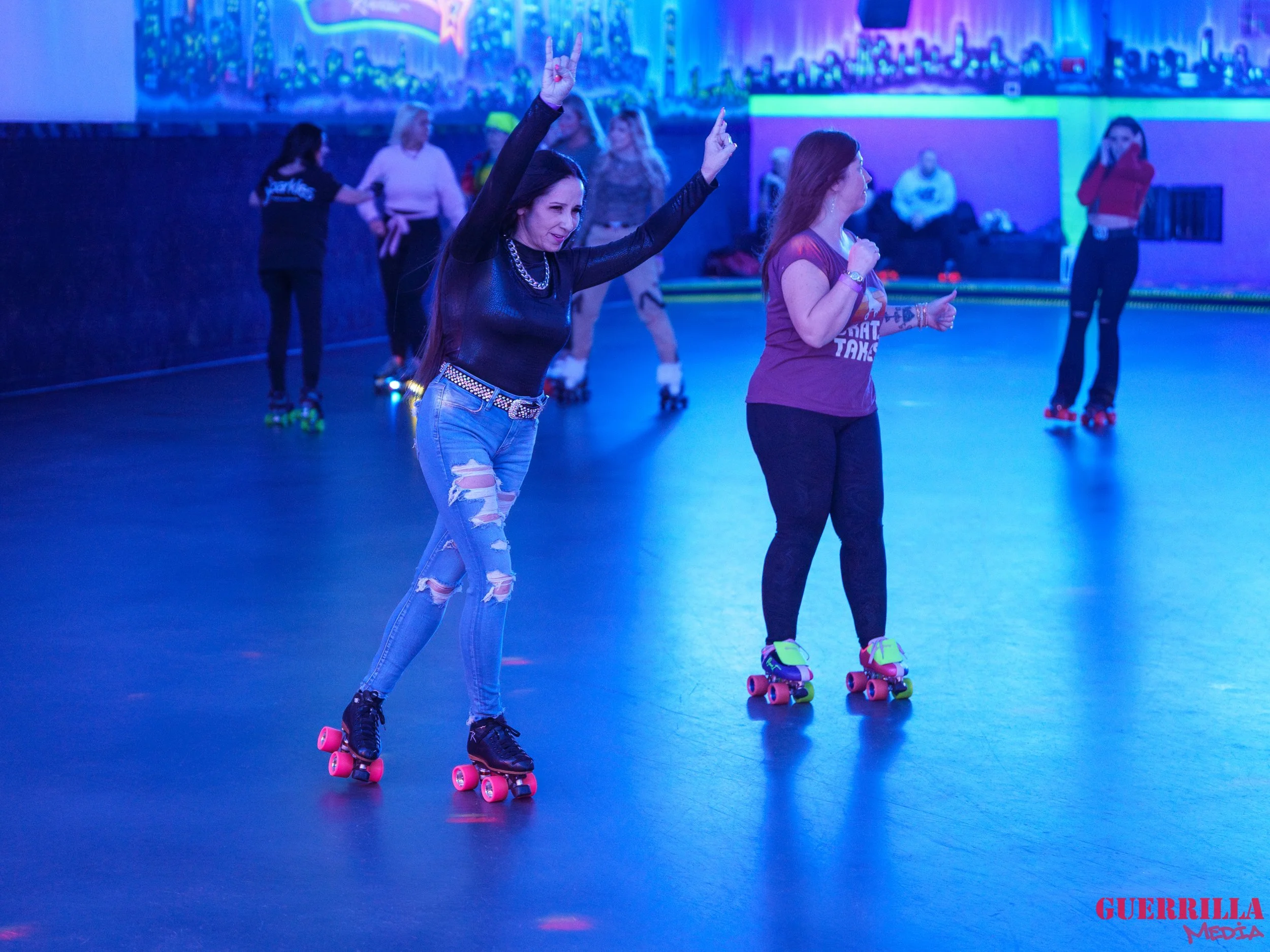 Women roller skating in a roller rink with vibrant lighting, some women are dancing, and others are skating. One woman in the foreground is making a peace sign with her hands.