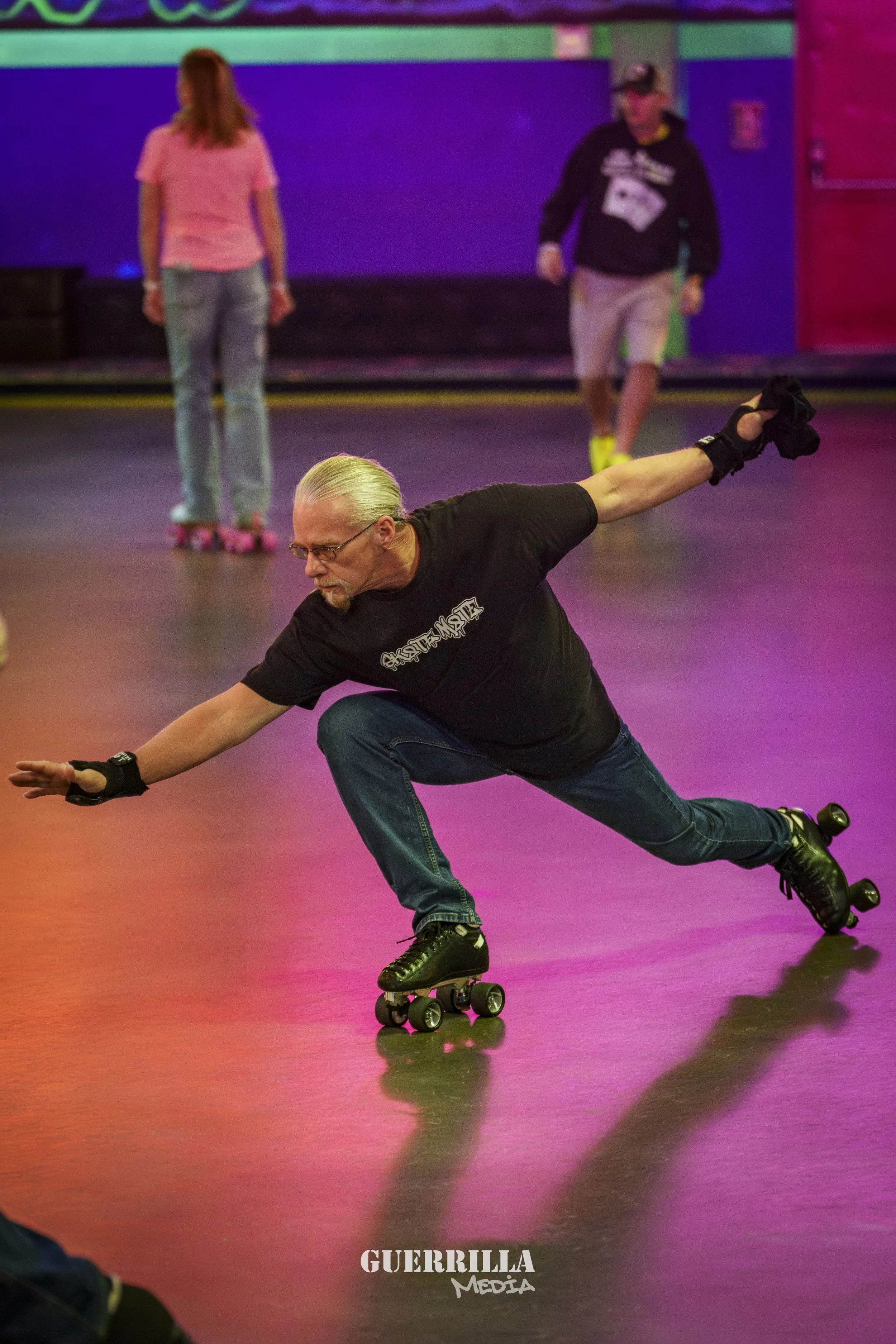 An older man skating on roller skates, balancing with arms outstretched in a pose, inside an indoor roller rink with colorful lighting, with two women in the background wearing roller skates.