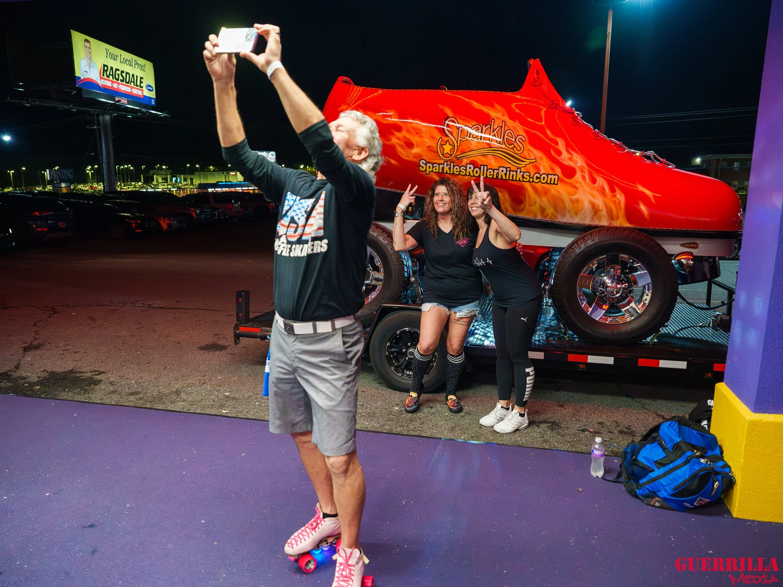 Two women posing for a photo at night in front of a large red roller rink shoe attraction. One woman is making a peace sign, and the other woman is also making a peace sign. A man is taking their photo with his phone, wearing a black shirt and gray s