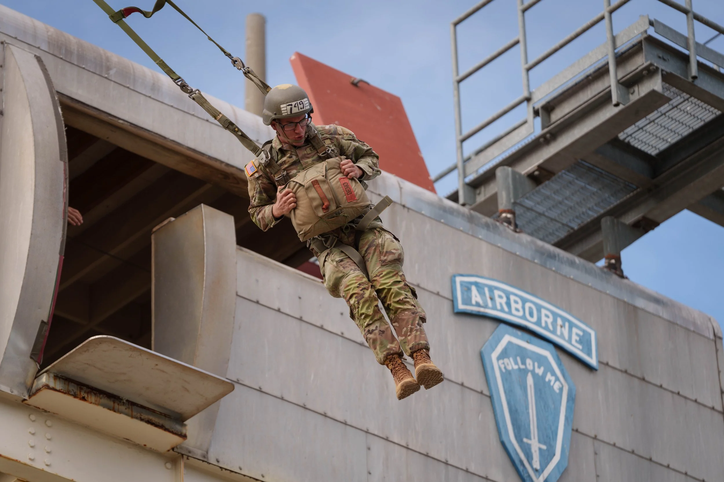 A soldier in camouflage uniform and helmet is descending on a harness from a building with military insignia, including a sign that says "AIRBORNE" and another that says "FOLLOW ME."