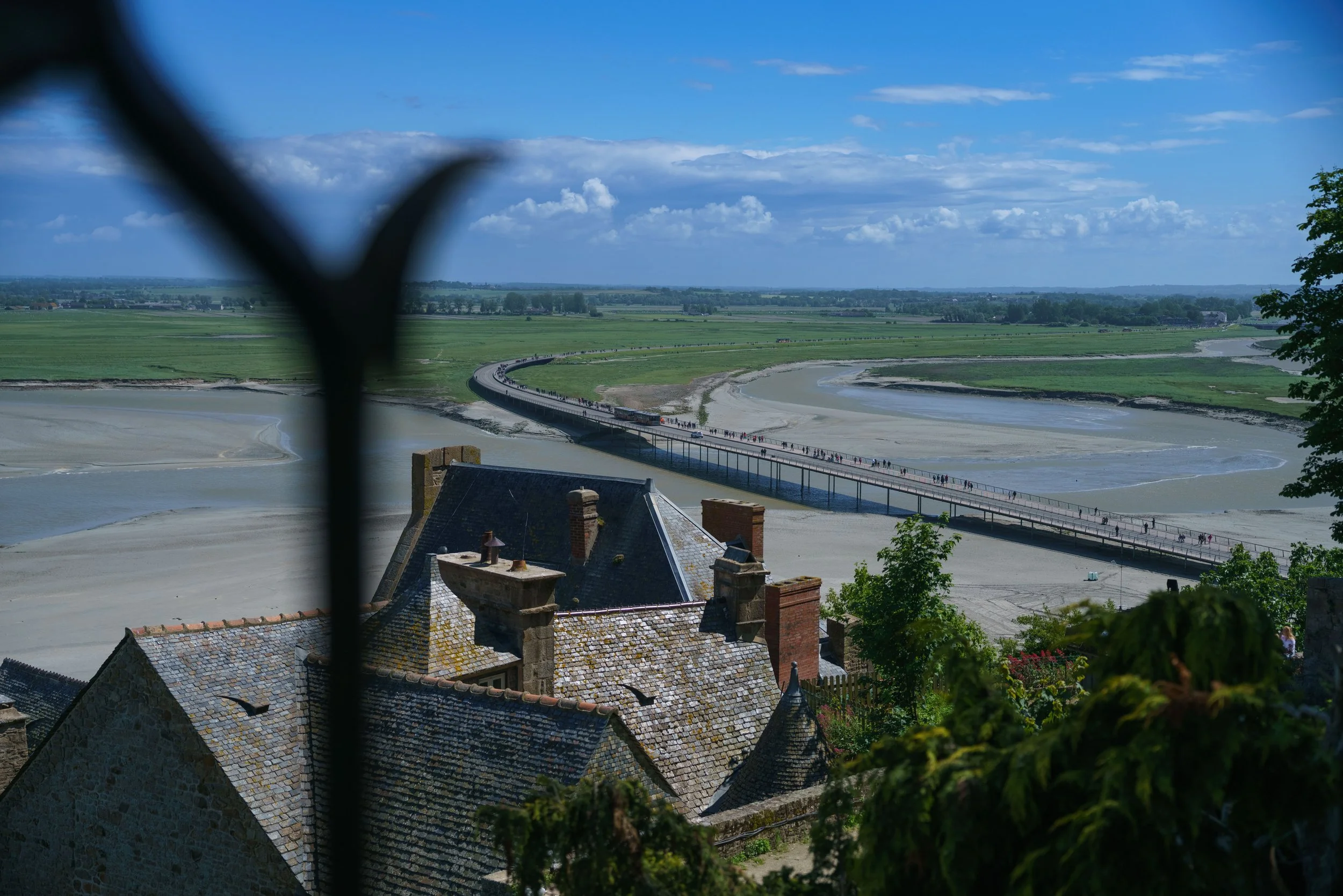 View of a long bridge over a river in a flat landscape, with green fields and a blue sky with clouds, from a high vantage point behind a building with steeply pitched roofs.
