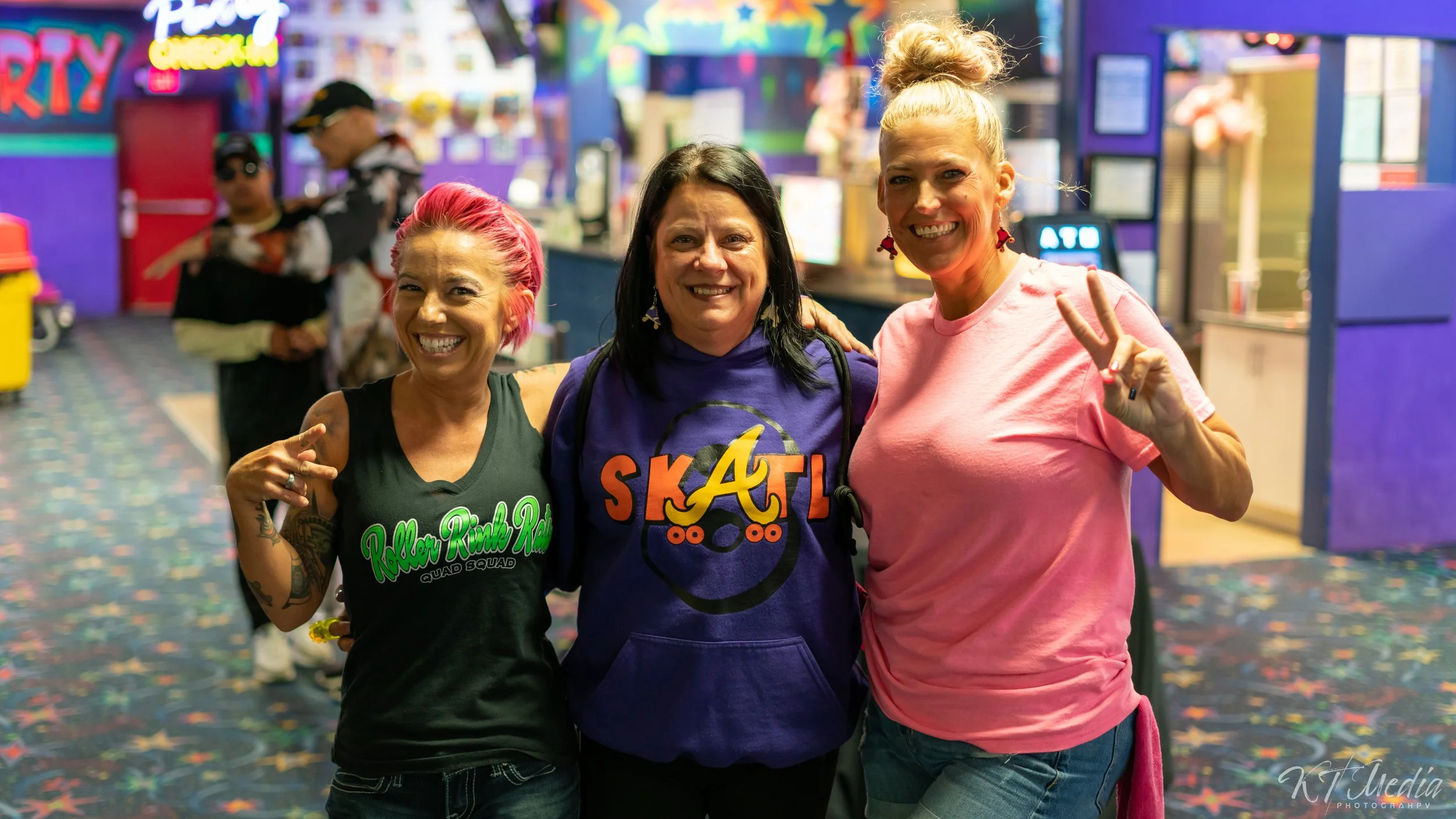 Three women smiling and posing for a photo at an indoor entertainment venue, with arcade games and colorful decorations in the background.