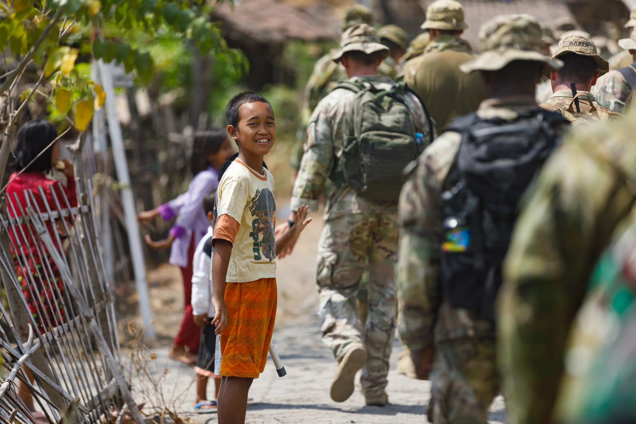 A young boy smiles and waves as soldiers walk past in a line; children stand behind a fence, watching.