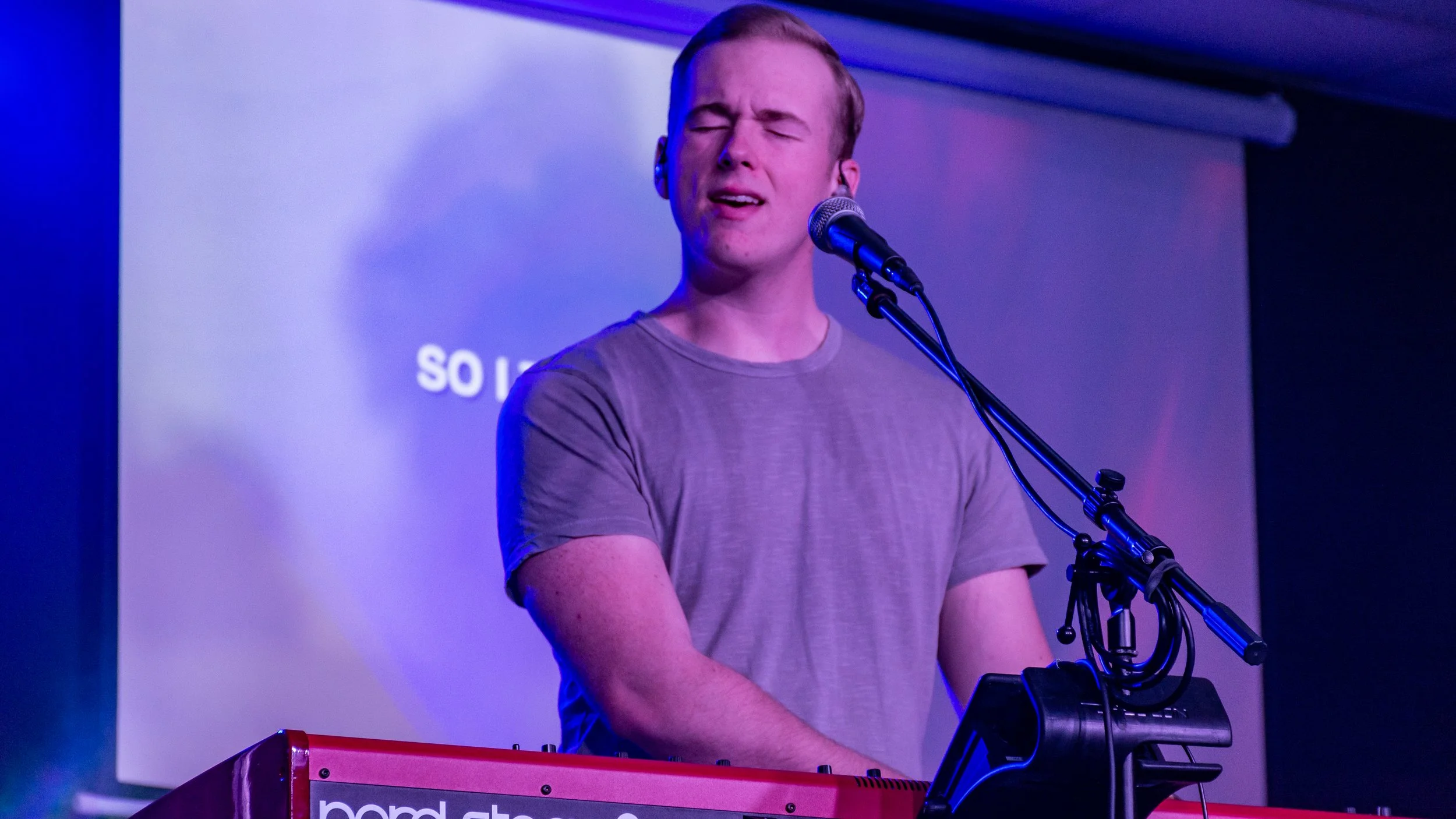 A man with closed eyes singing into a microphone while playing a red keyboard on stage, in front of a white screen with text.