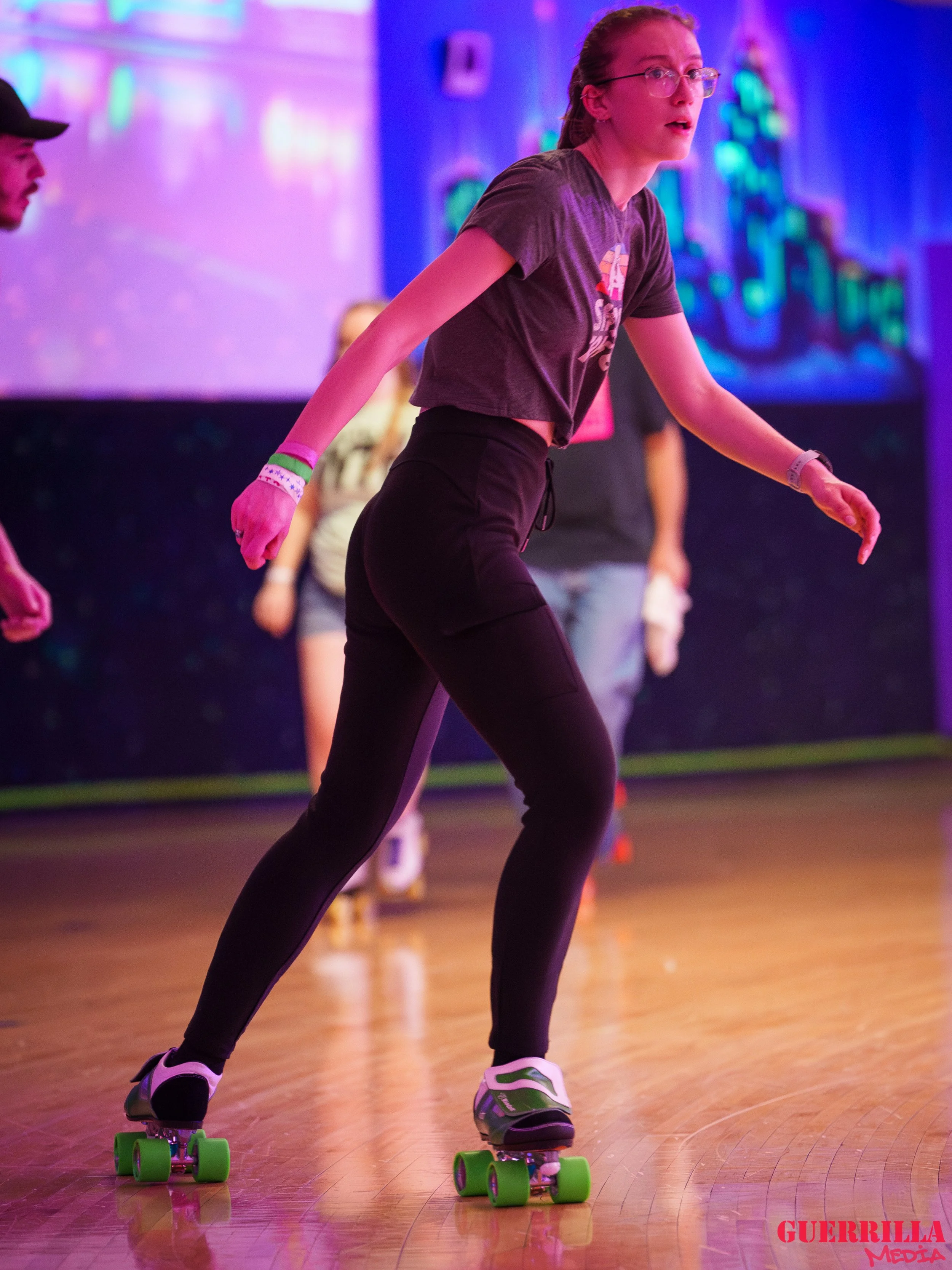 A young woman wearing glasses, a dark t-shirt, and black pants is roller skating indoors on a wooden floor with colorful neon lighting. She appears to be balancing or practicing, with other people visible in the background.