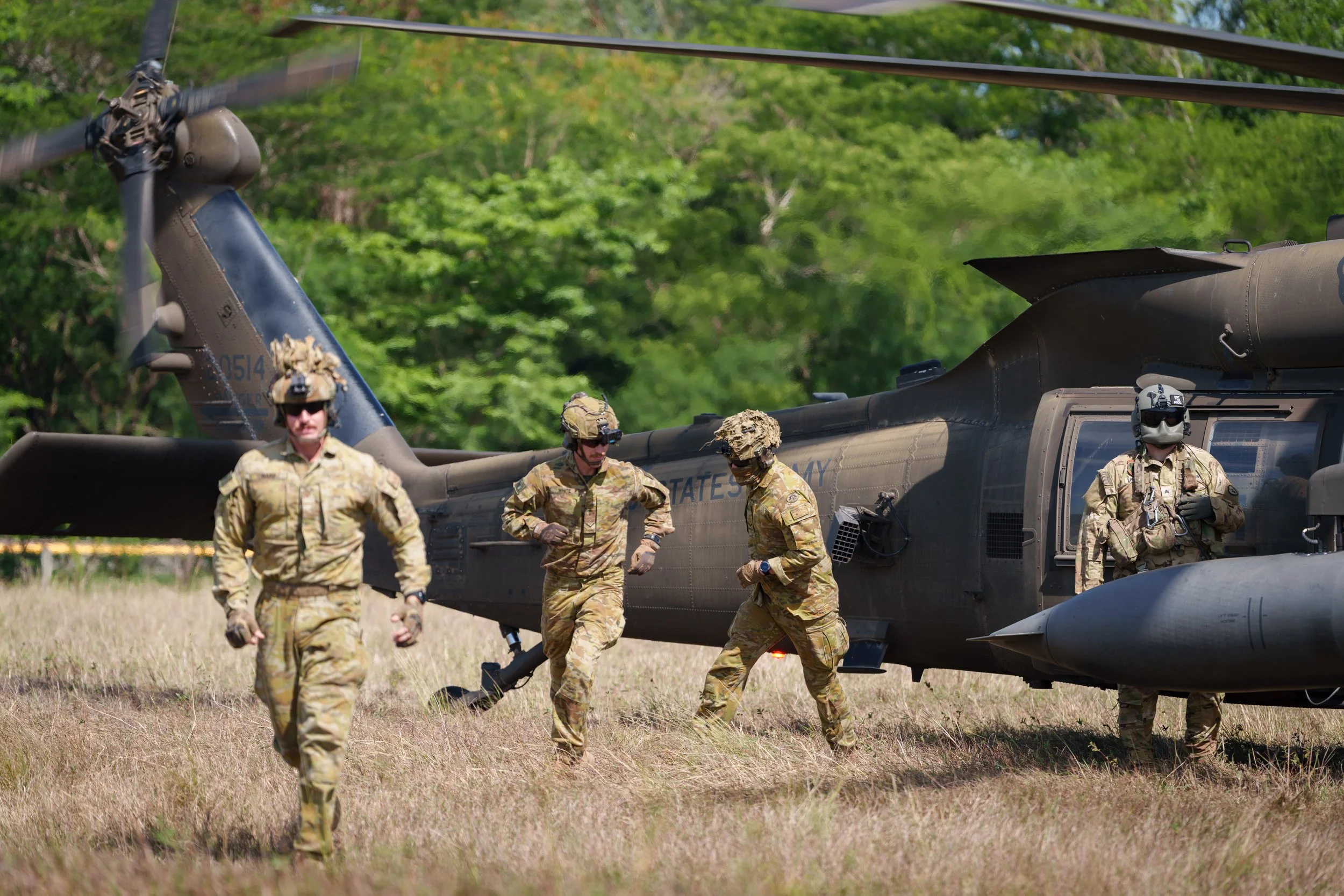 Four military personnel in camouflage uniforms and helmets near a black helicopter on a grassy field with green trees in the background.