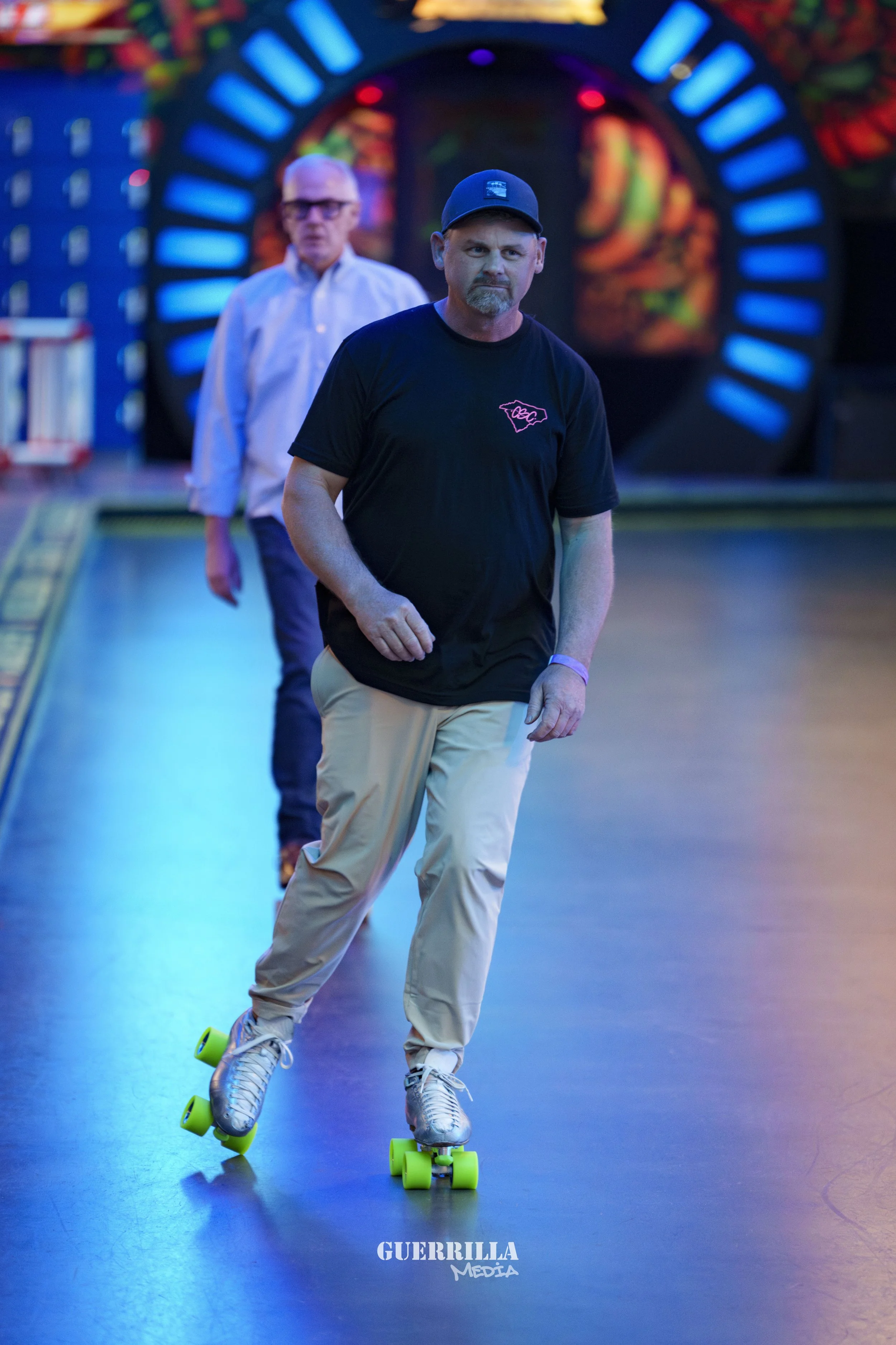 A man roller skating on an indoor rink with colorful neon lights in the background, wearing a black T-shirt, beige pants, a black cap, and gray skate shoes with green wheels. Another man in a white shirt and glasses is in the background.