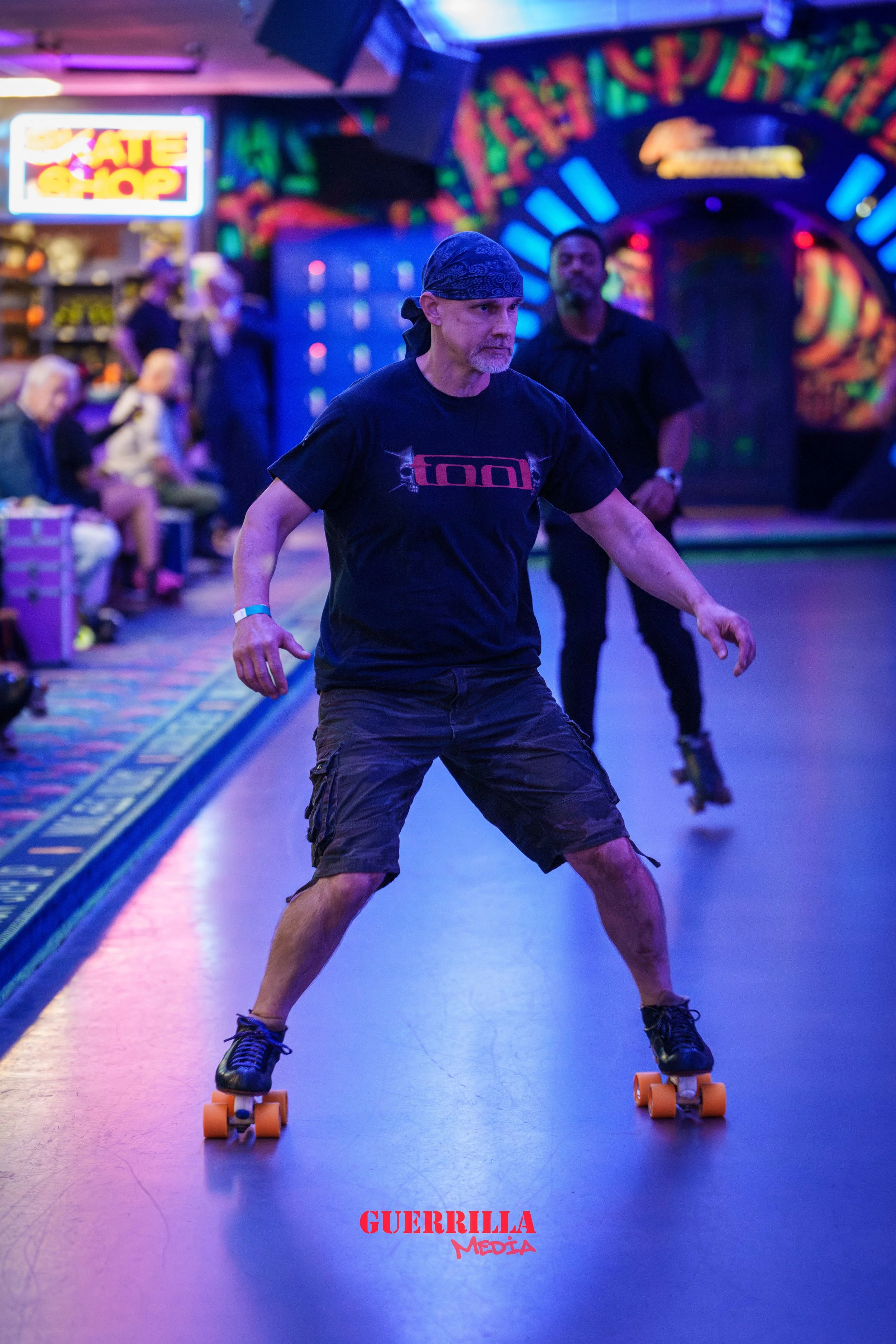 A man roller skating indoors at a colorful rink, wearing a black bandana and T-shirt, with spectators watching in the background.