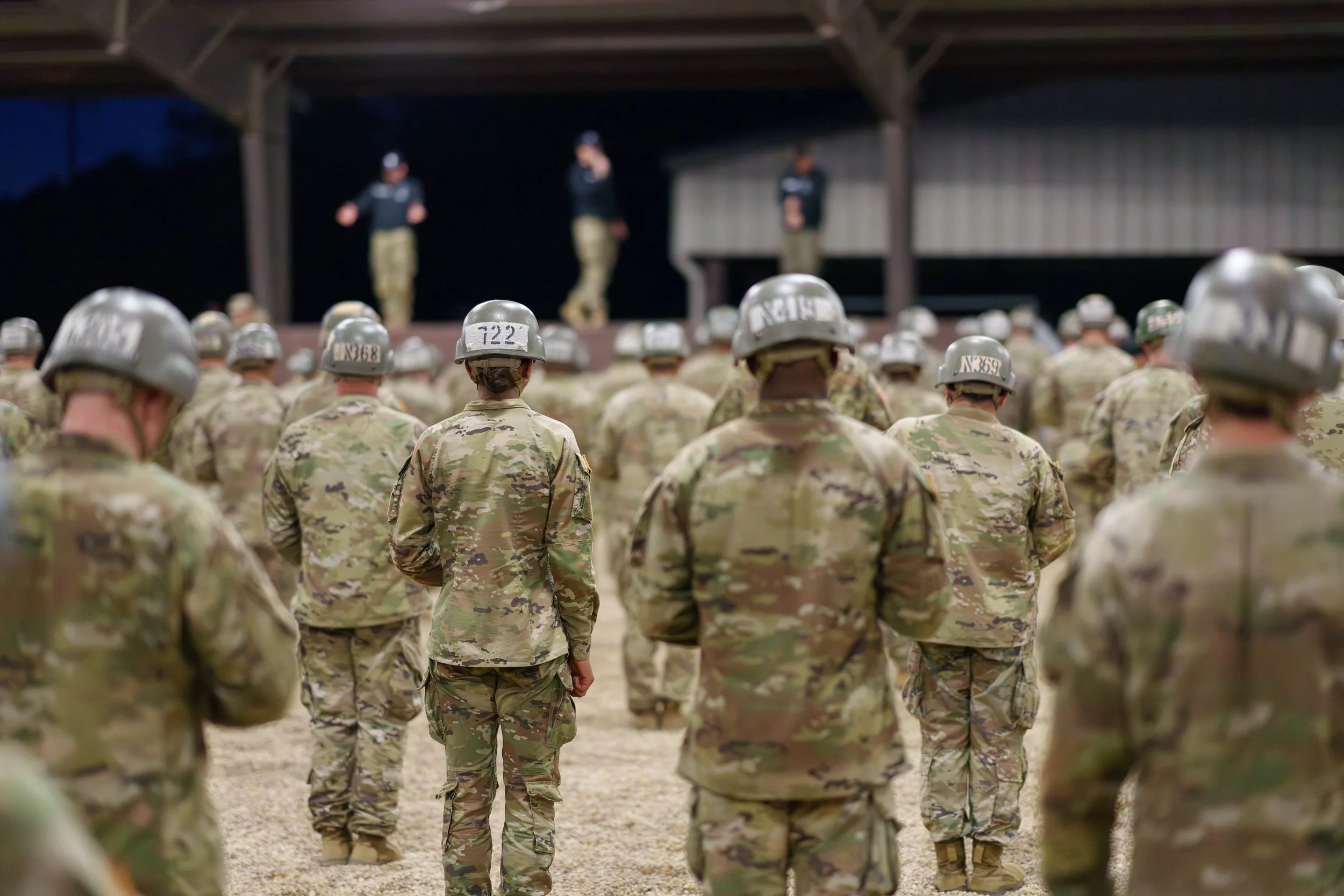 Military soldiers in camouflage uniform and helmets standing in formation at night, with others standing on a stage in the background.