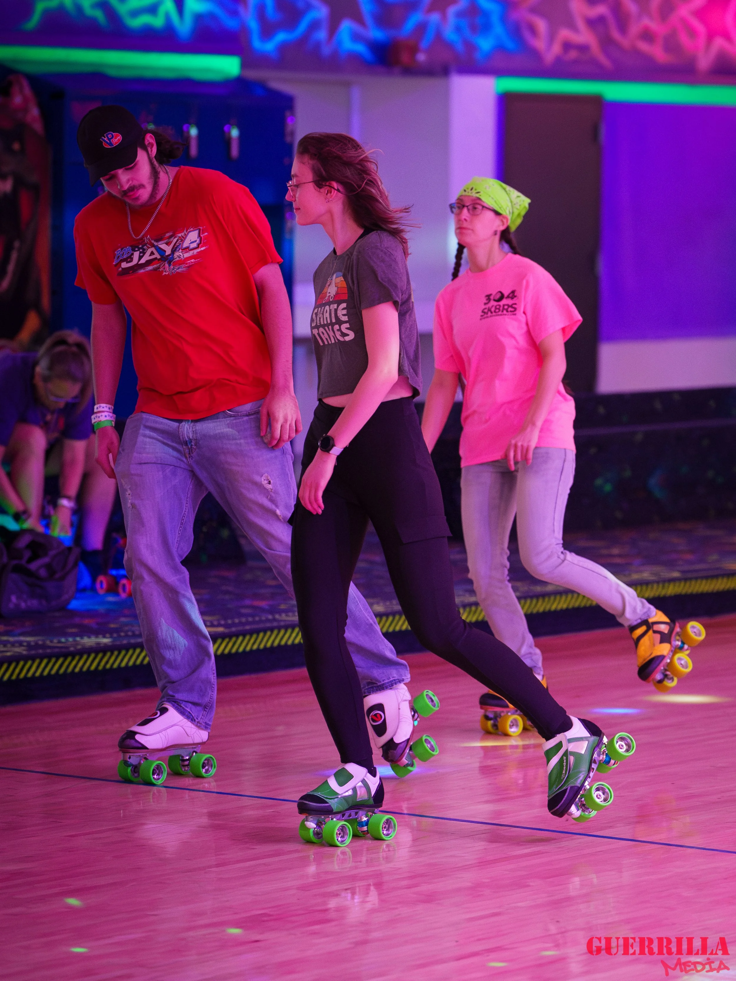 Three people roller skating at an indoor skating rink with neon lights. Two women and one man, all wearing casual clothing and roller skate shoes, are skating together in a line. The background has colorful glow decorations and others skating or sitt