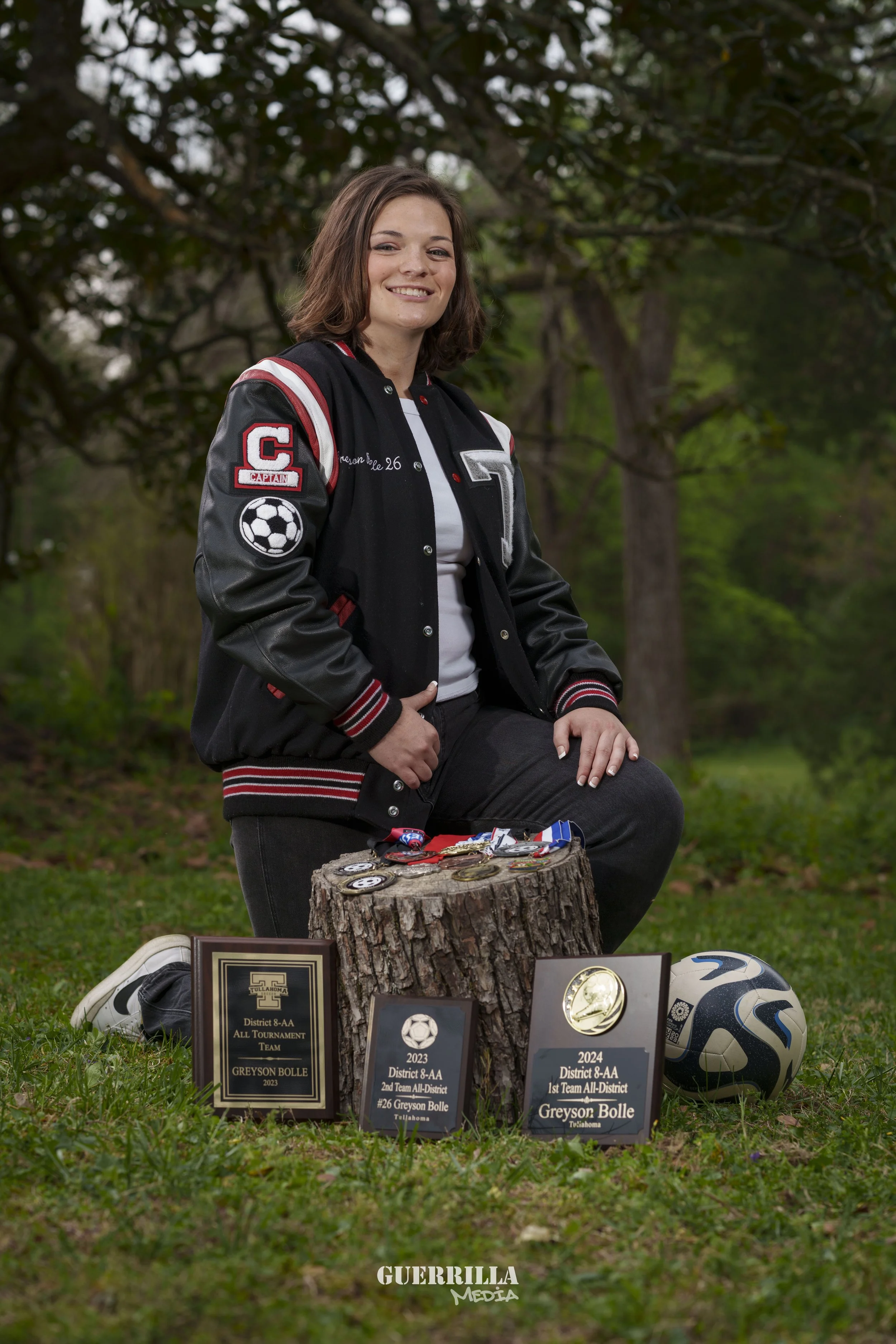 A young woman with shoulder-length brown hair, dressed in a black varsity jacket with red and white accents, kneeling on one knee next to a tree stump with medals and trophies displayed on it. She is outdoors in a park-like setting with trees and gre