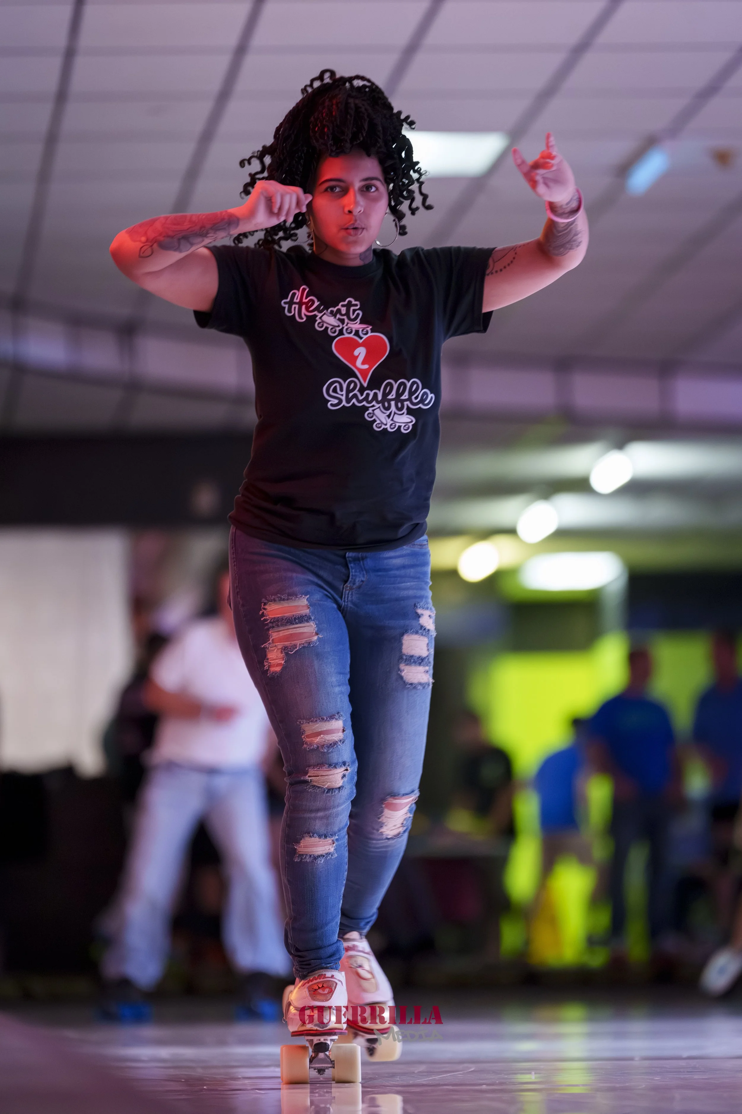 A woman roller skating indoors, wearing a black T-shirt with the text 'Heart to Shuffle' and ripped jeans.