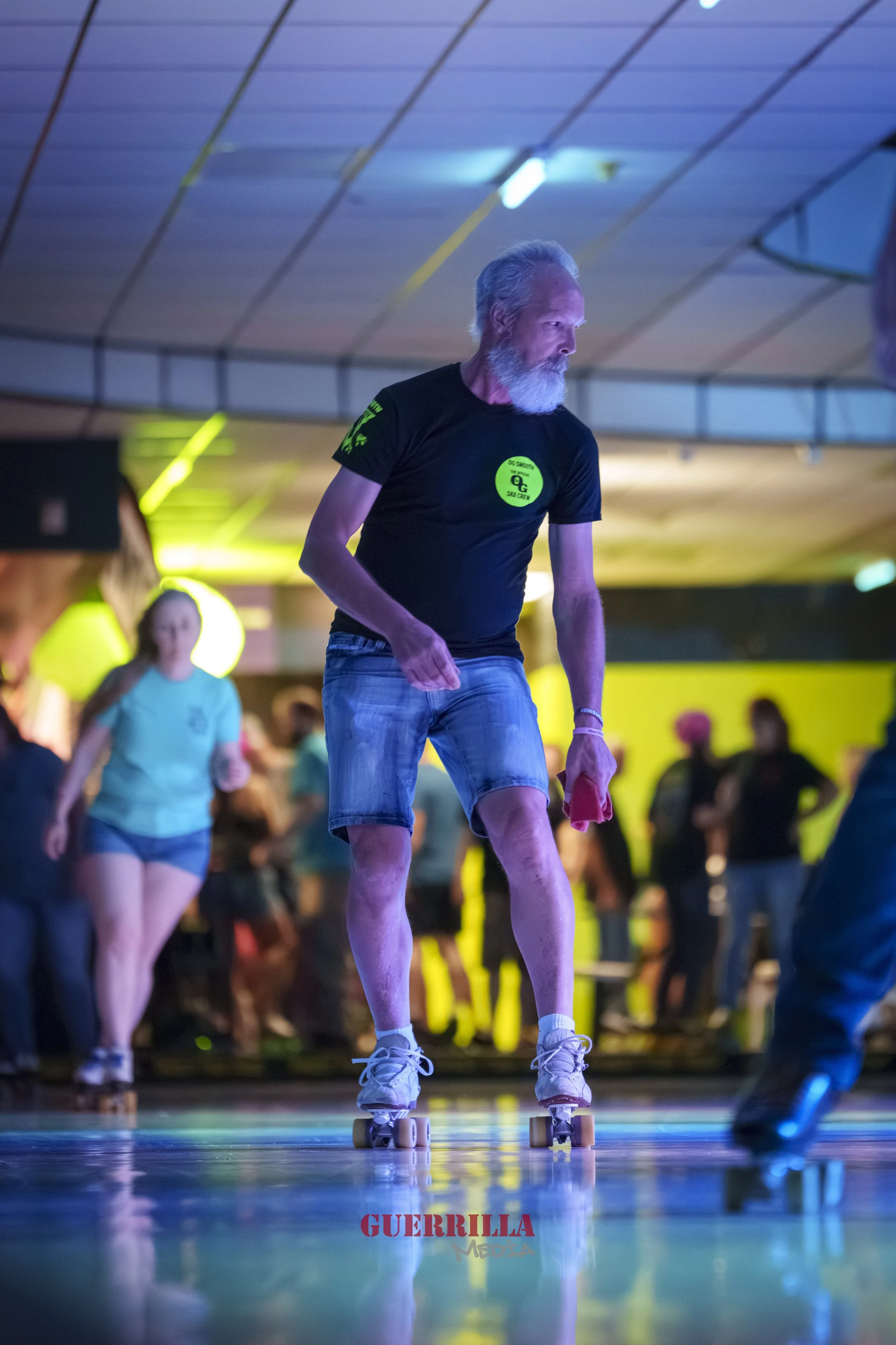 A man roller skating indoors with a crowd in the background, colorful lighting, and a watermark reading 'GUERRILLA MEDIA' at the bottom.