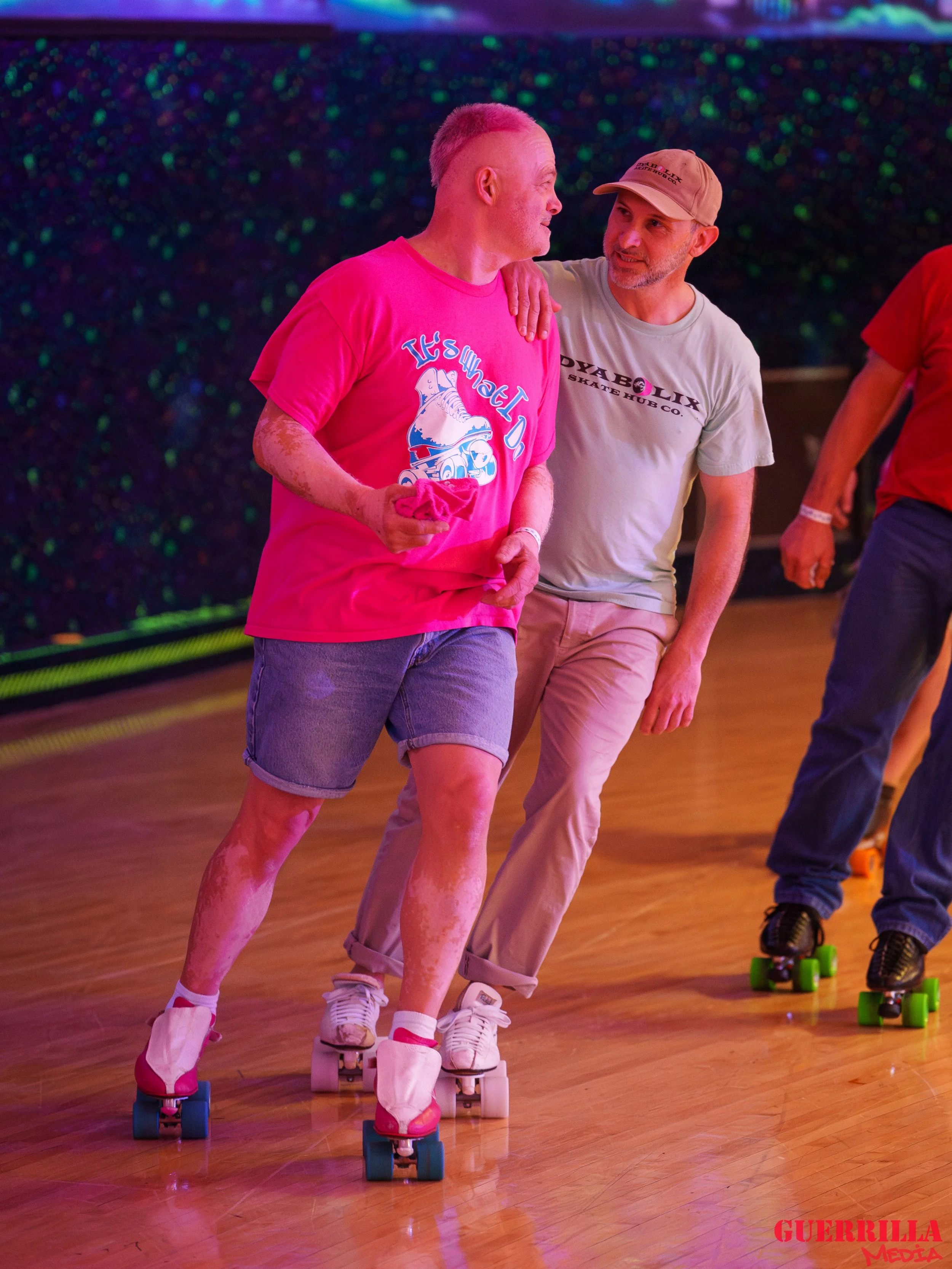 Two men roller skating inside a rink, one in a pink T-shirt and the other in a gray T-shirt, sharing a moment and smiling at each other.