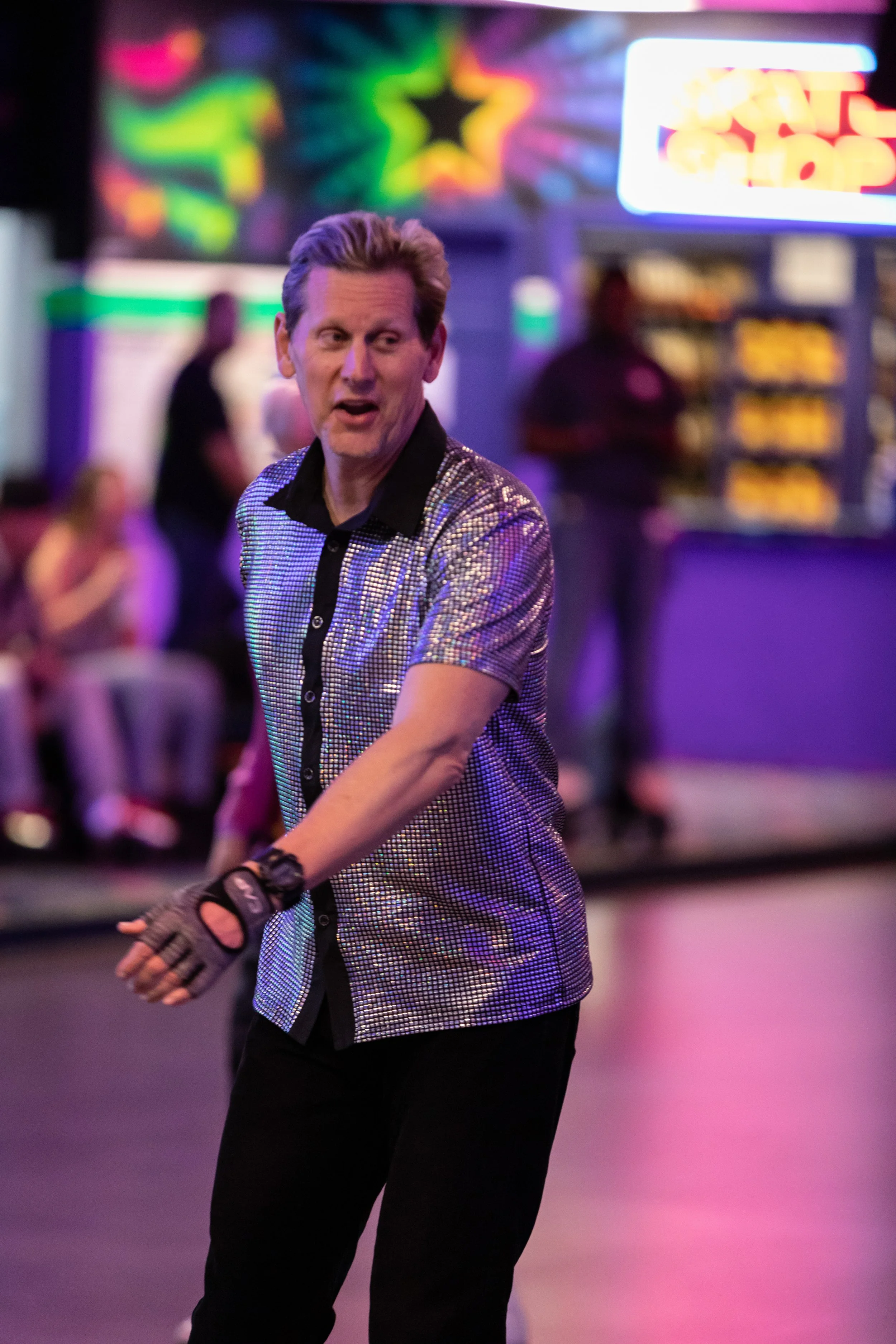 A man wearing a shiny, metallic, short-sleeve shirt and fingerless gloves roller skating in an indoor skating rink.