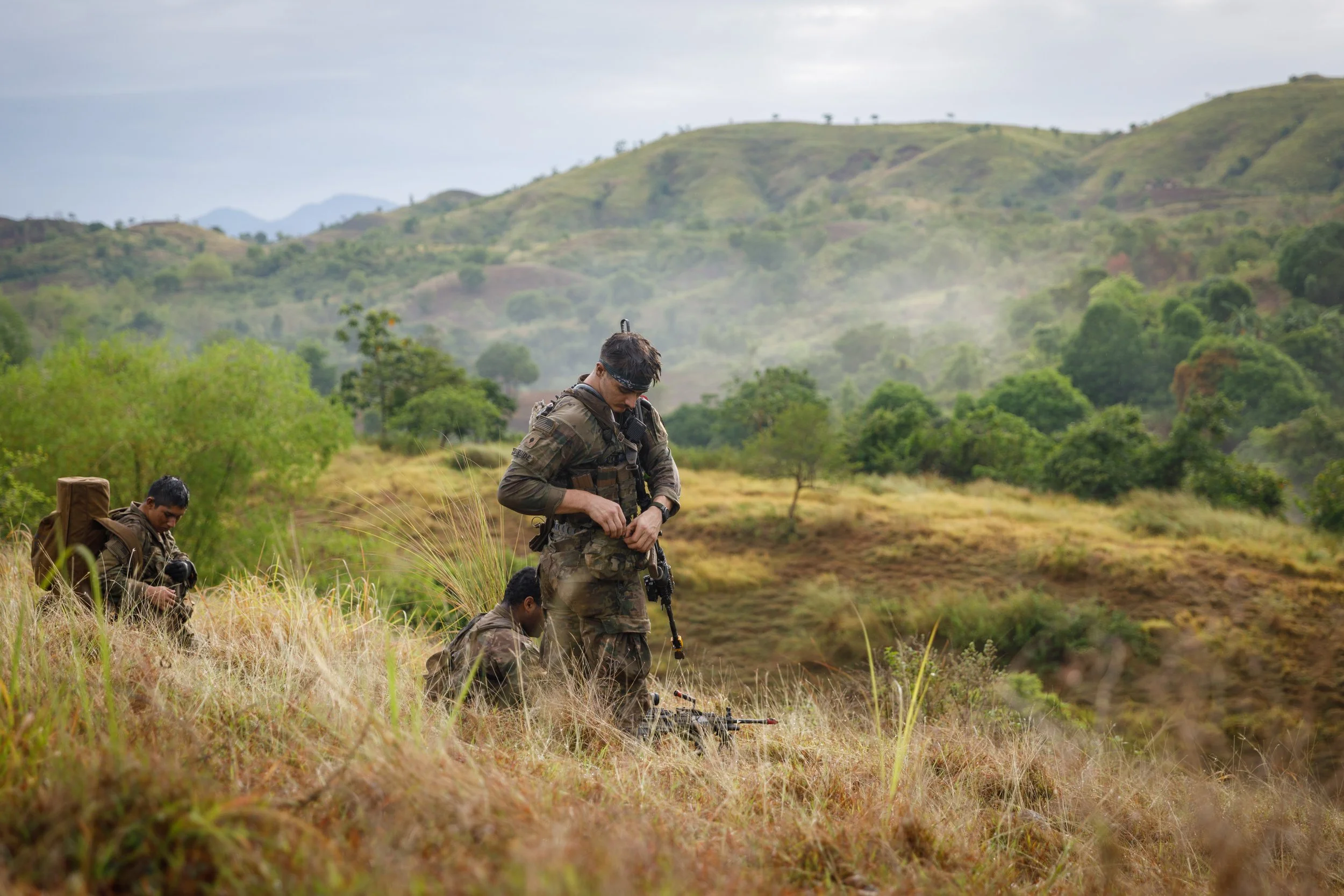 Four soldiers in camouflage uniforms and gear, one kneeling, two standing, in a grassy, hilly landscape with trees and mountains in the background.