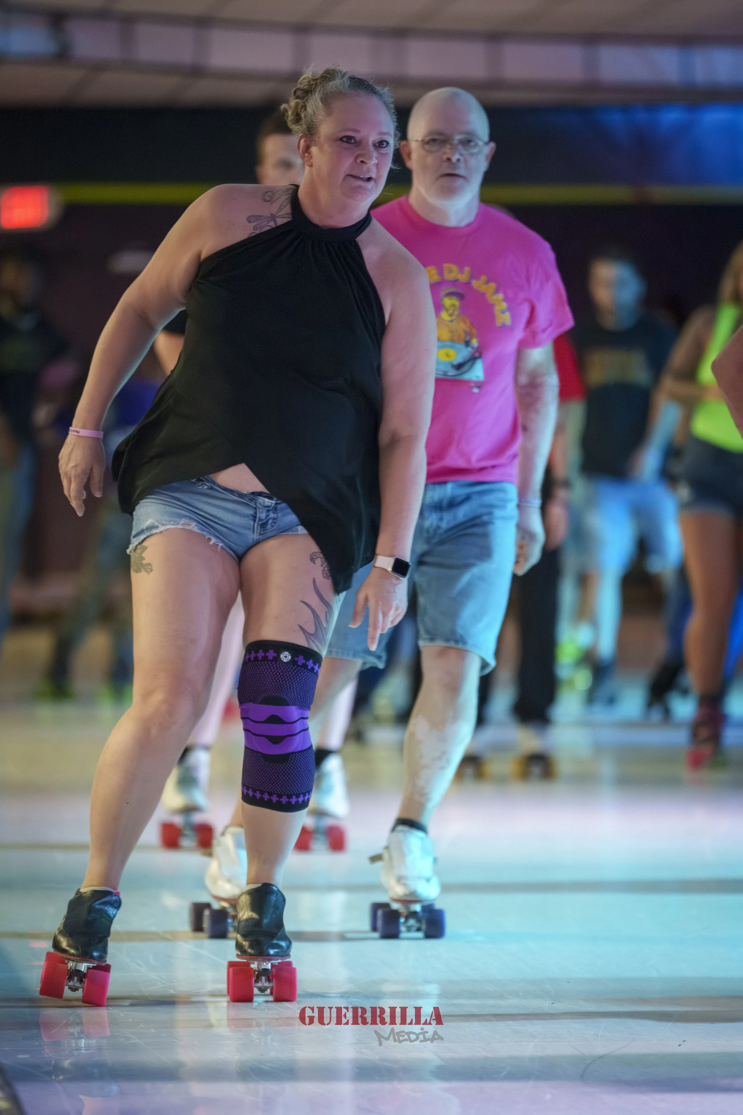 People roller skating indoors, in a line, with a woman in the foreground wearing a black halter top, denim shorts, black shoes, and a purple leg brace, leading the group.