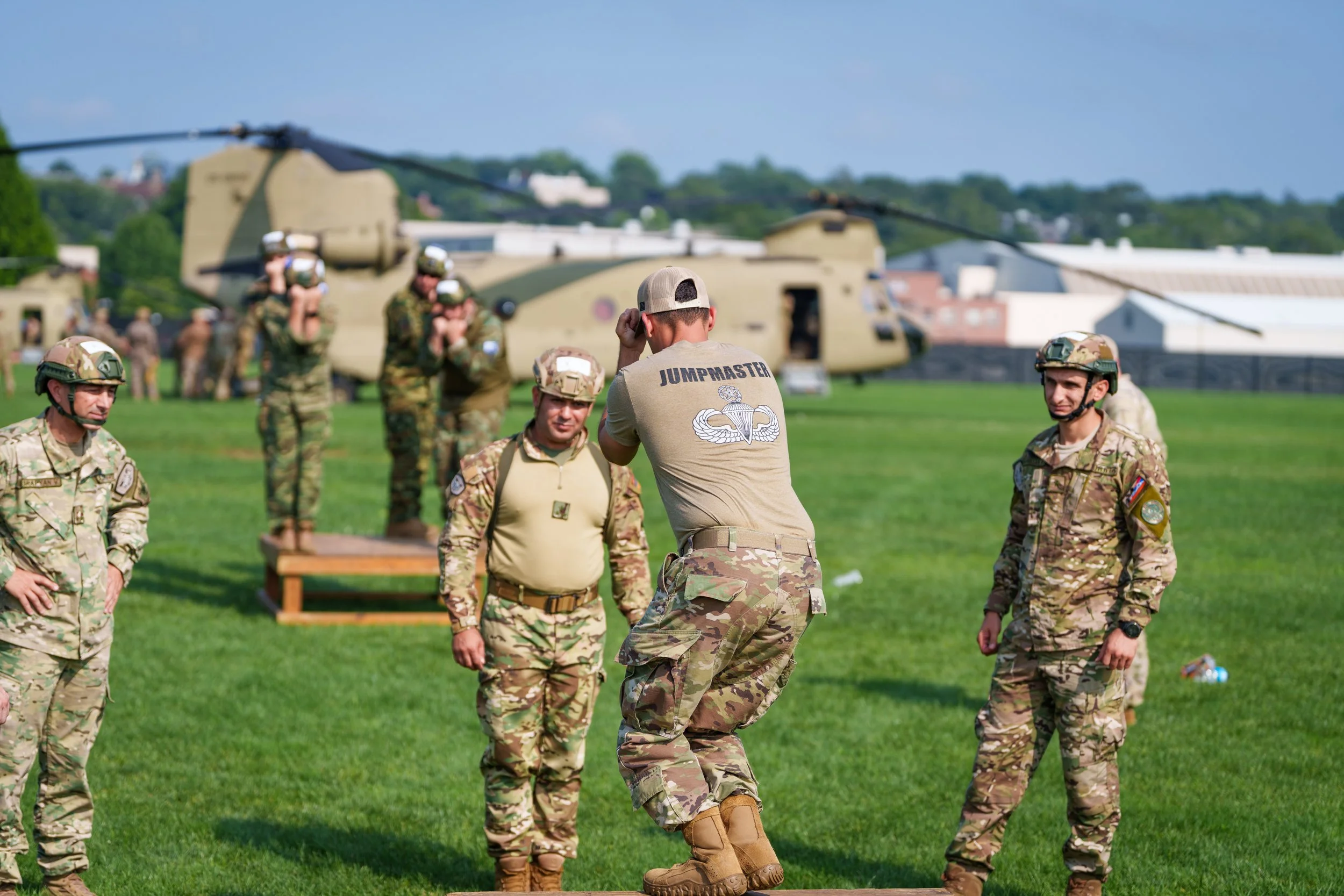 Military personnel in camouflage uniforms participating in a training exercise on a grassy field with a helicopter in the background.