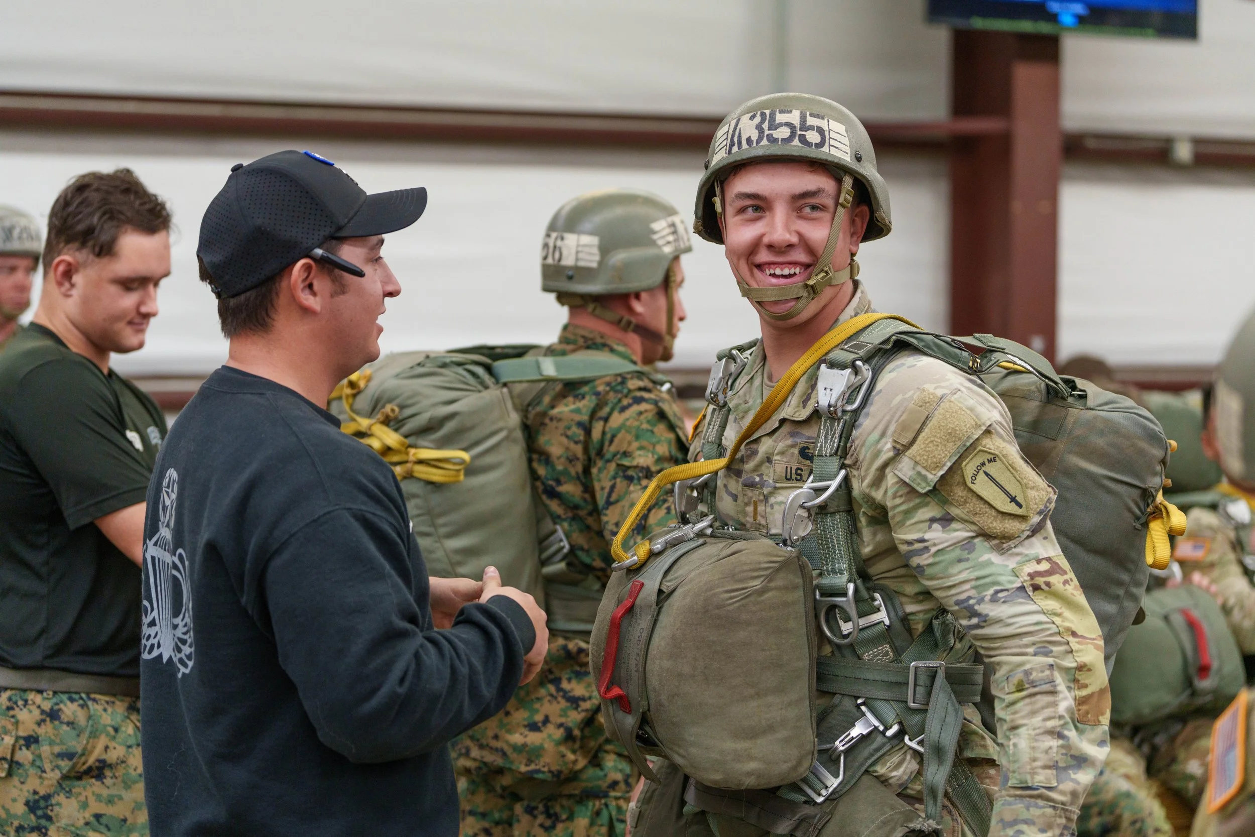 A smiling soldier in camouflage uniform and helmet standing with others in military gear, inside a hangar or large indoor facility.