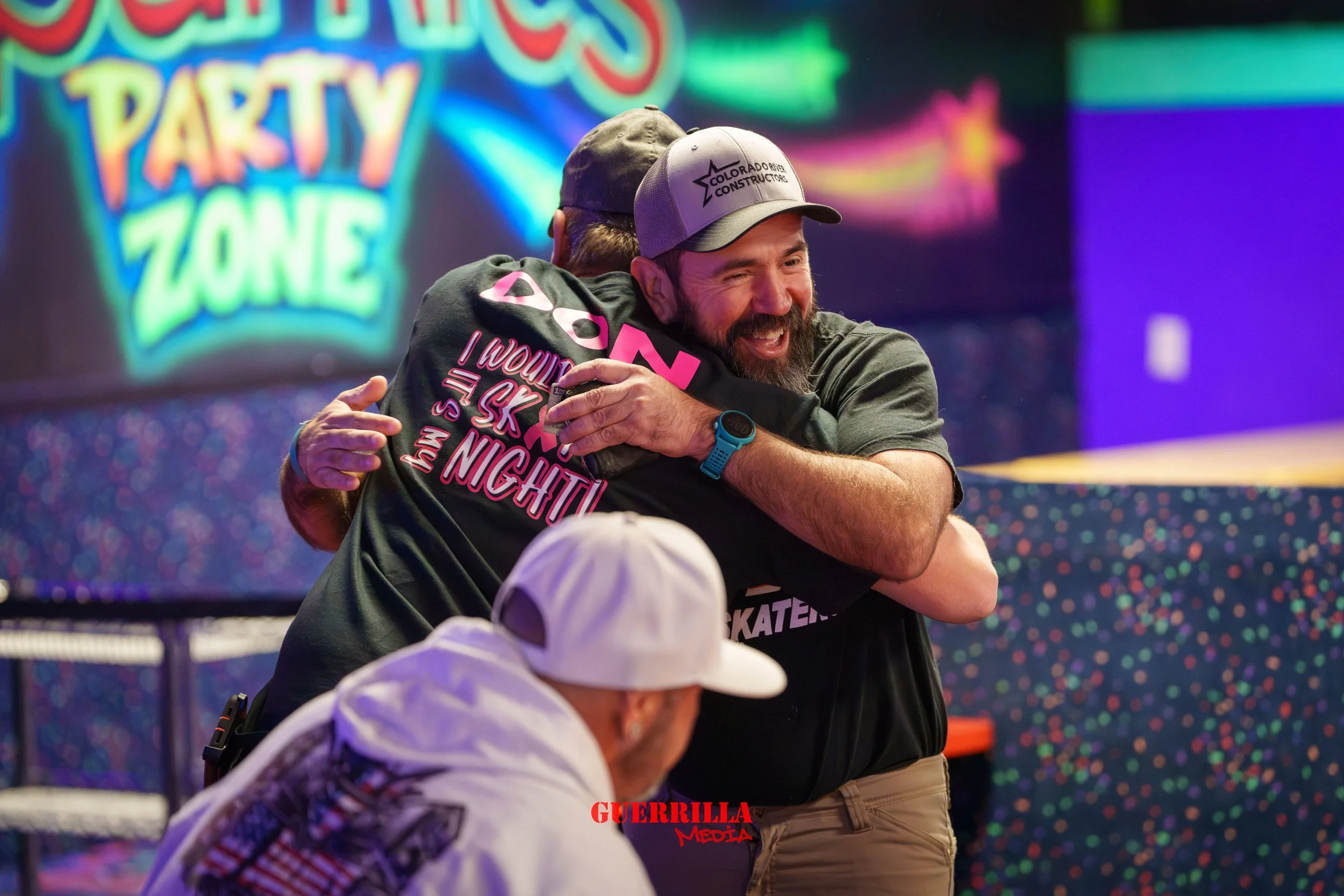Two men hug and smile at a roller skating rink with colorful neon lights in the background. One wears a black shirt and a gray hat, and the other wears a white cap and a white hoodie.