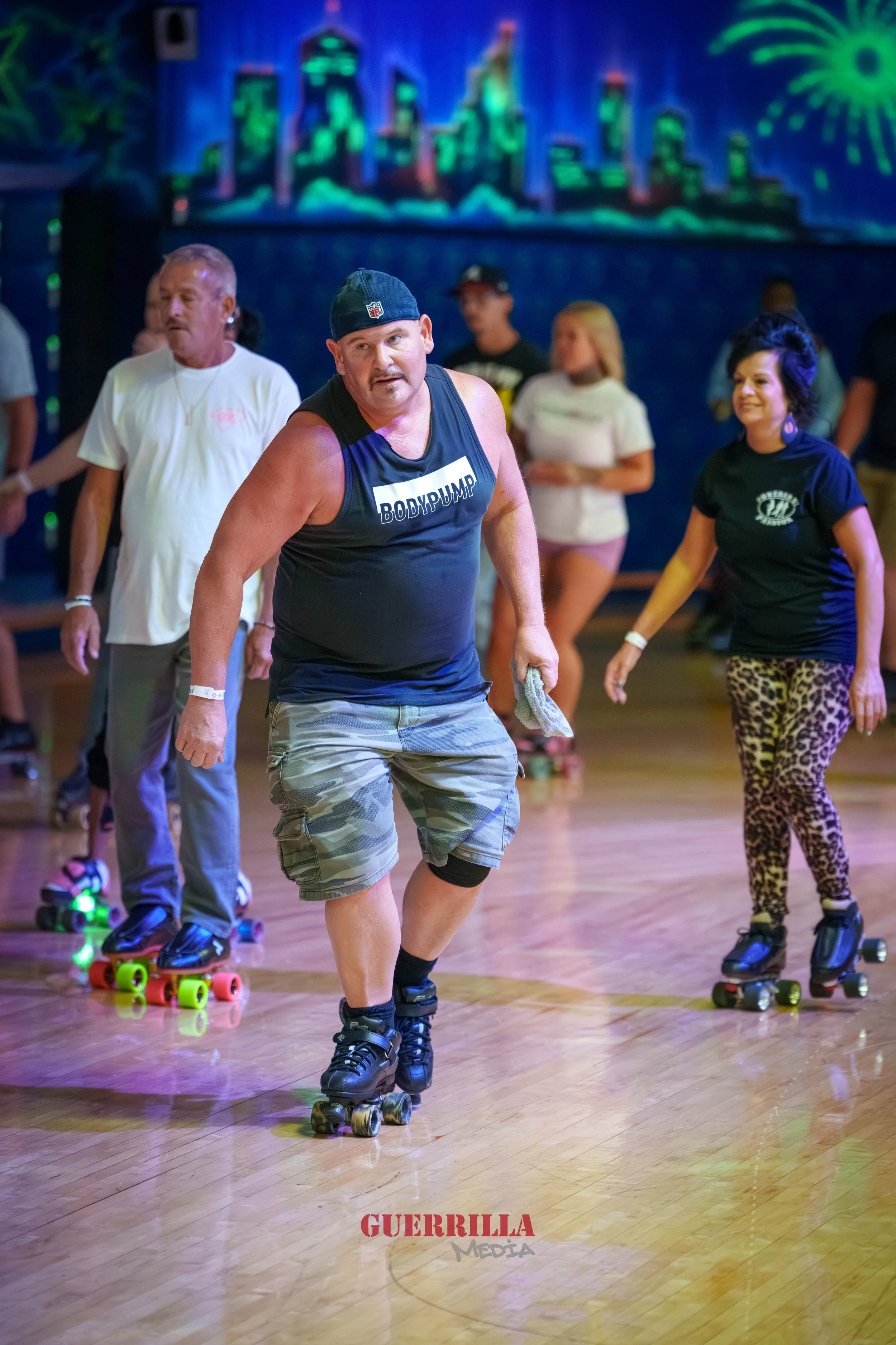 People roller skating indoors in a neon-lit skate rink with a cityscape mural and fireworks on the wall.