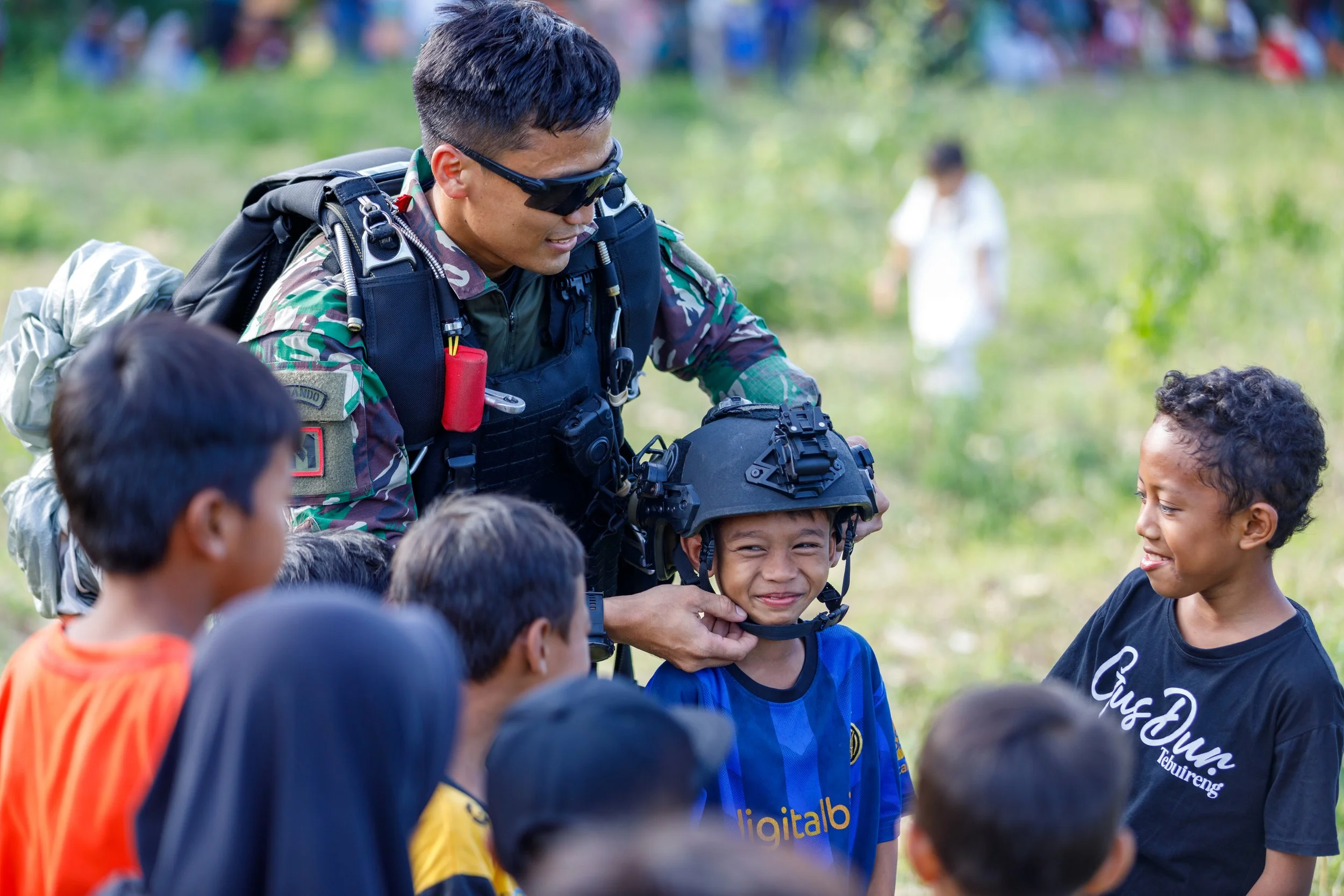 A soldier in camouflage uniform and tactical gear smiling and helping a young boy put on a military helmet during an outdoor event with children surrounding them.