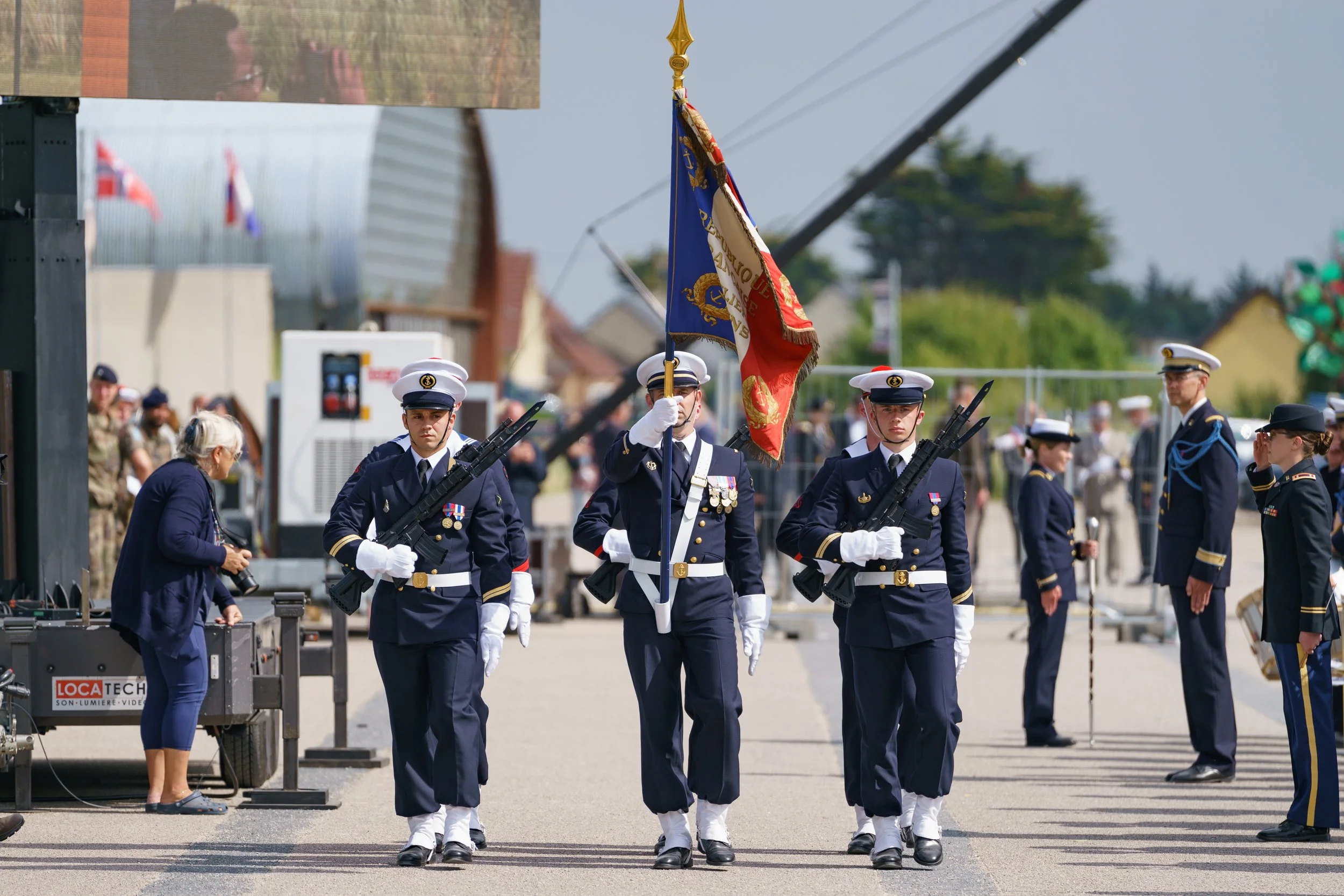 Military parade with soldiers in formal dress uniforms marching, one carrying a flag, during a public event.