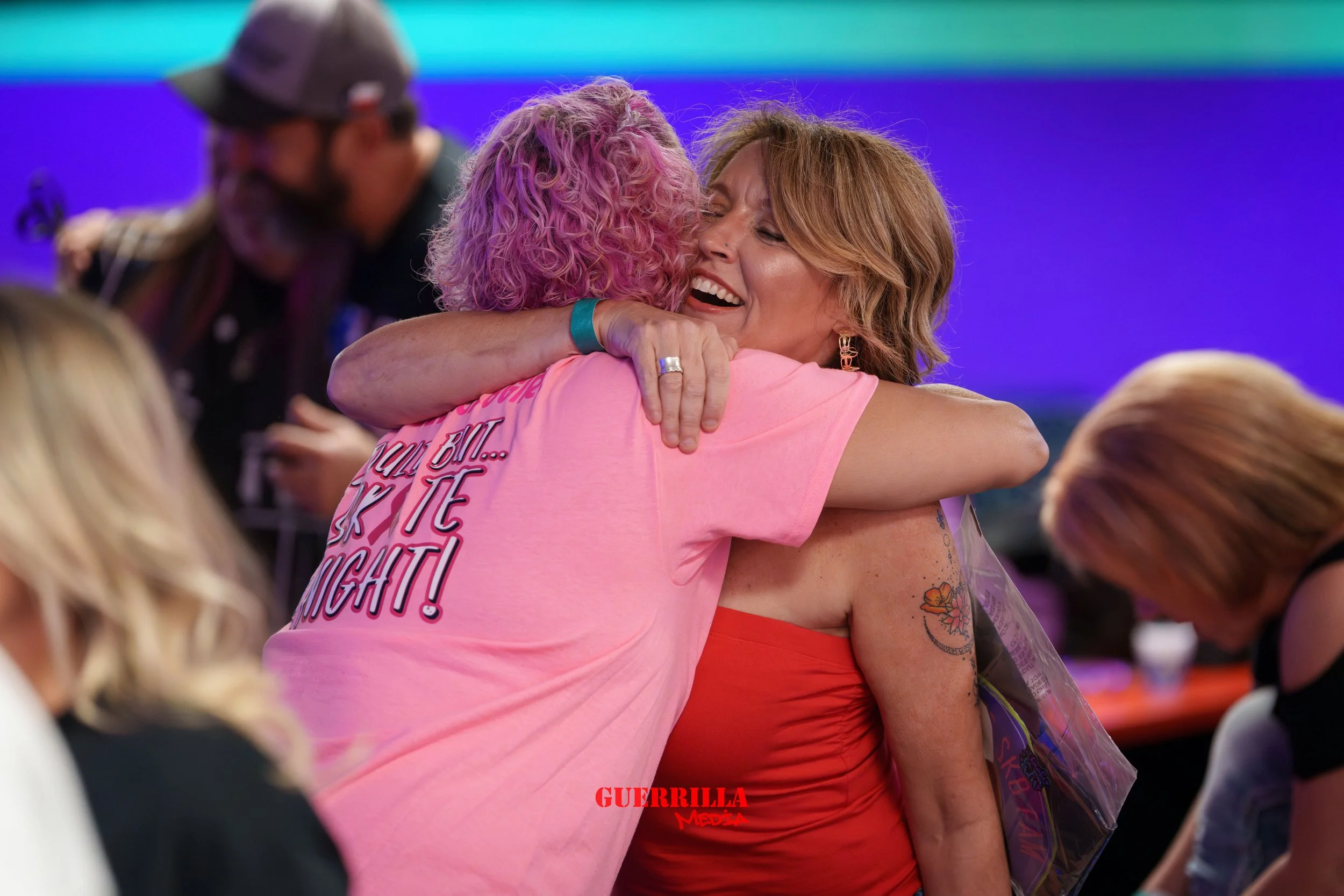 Two women hugging warmly, smiling, with one wearing a pink shirt and the other in a red dress. The background is colorful and blurred, indicating an indoor event.