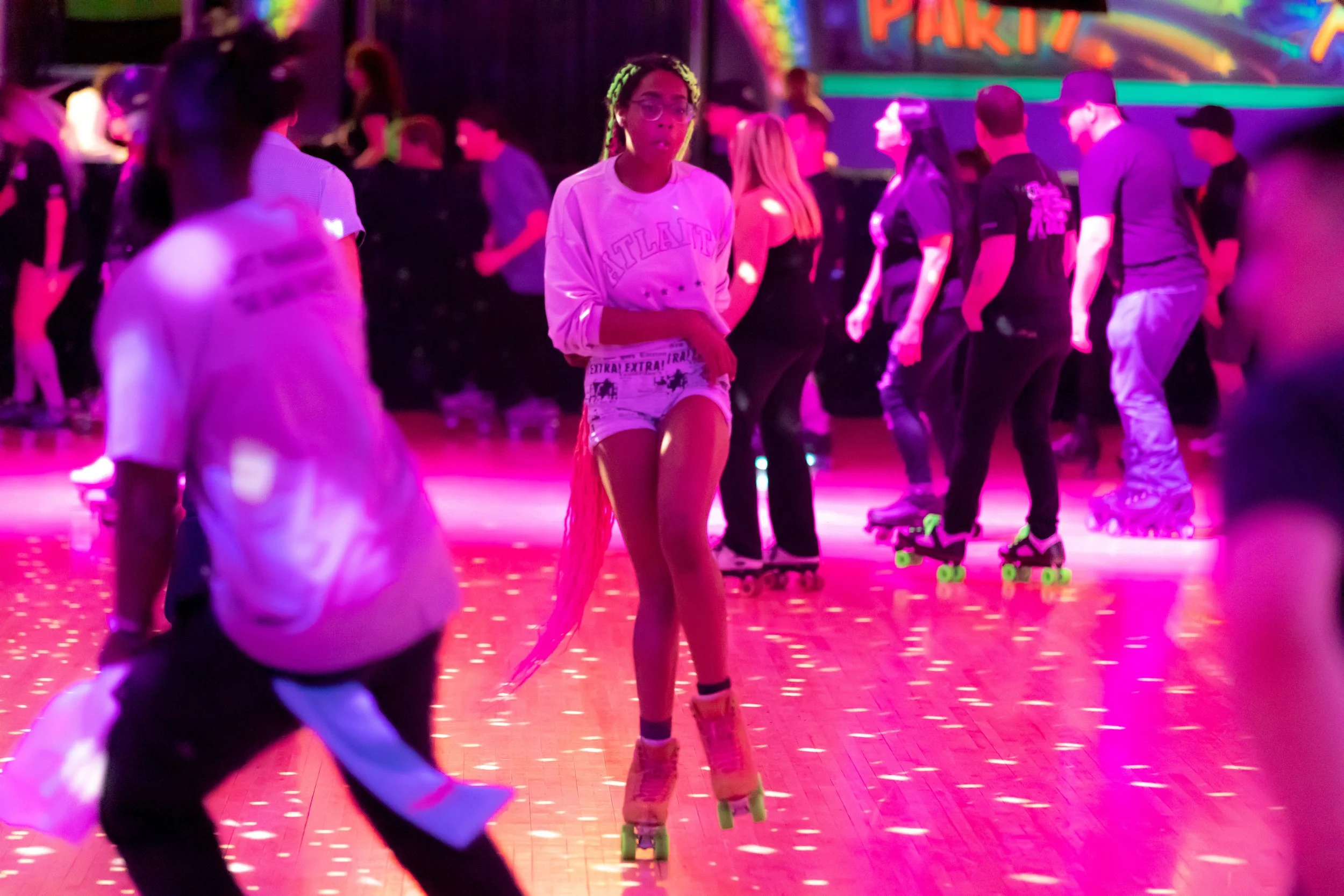 People roller skating at a party rink with colorful neon lighting and a 'Party' sign in the background.