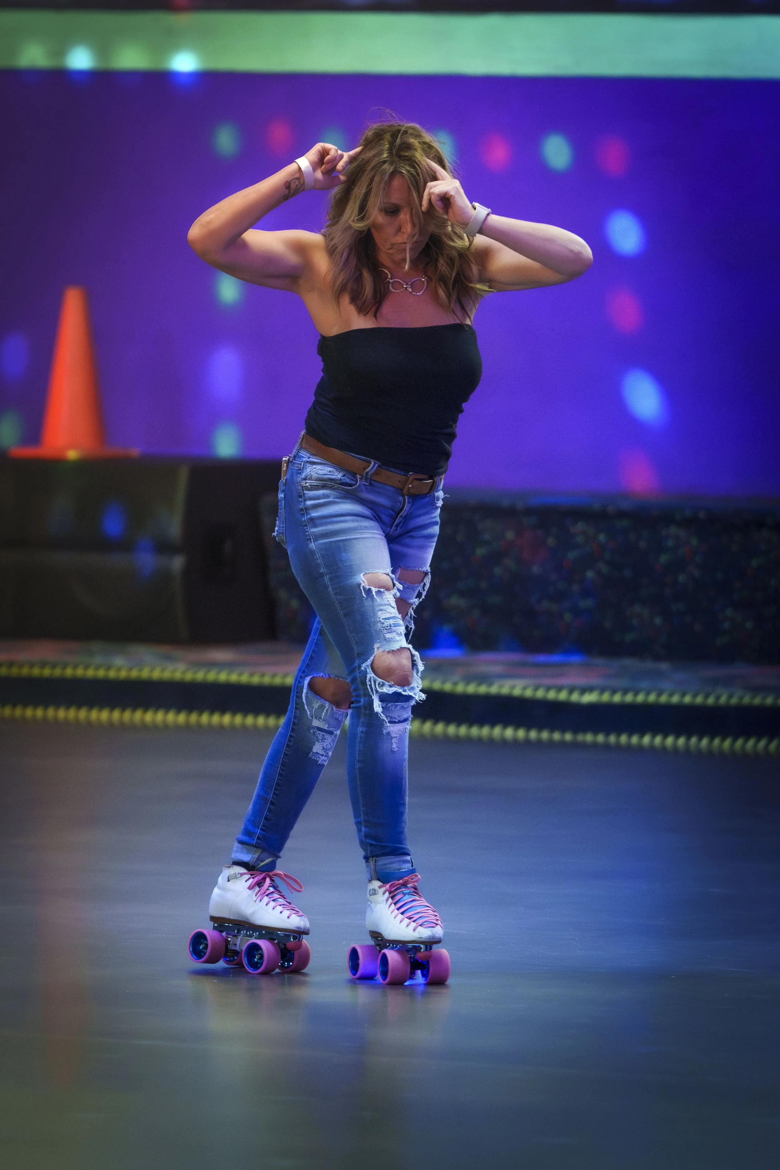 A woman roller skating indoors wearing a black tube top, ripped jeans, pink laced roller skates, and a necklace, with colorful lights in the background.