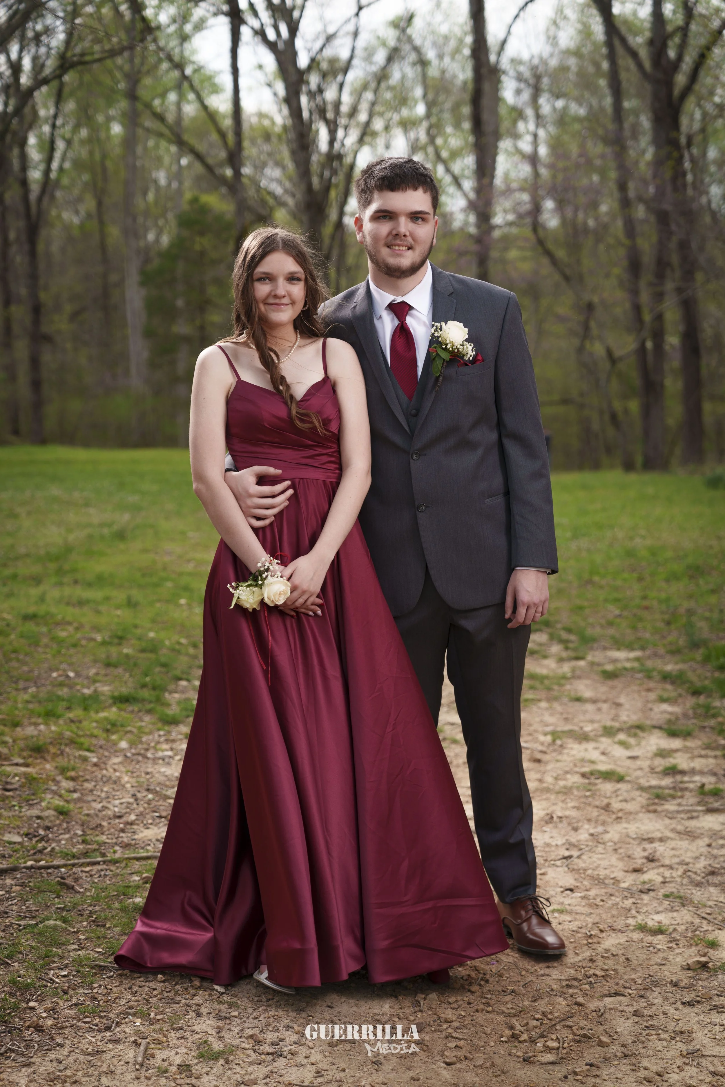 A young couple dressed in formal attire, standing outdoors on a dirt path with green grass and trees in the background. The girl wears a burgundy gown and holds a white flower, while the boy wears a dark suit with a red tie and a boutonniere.