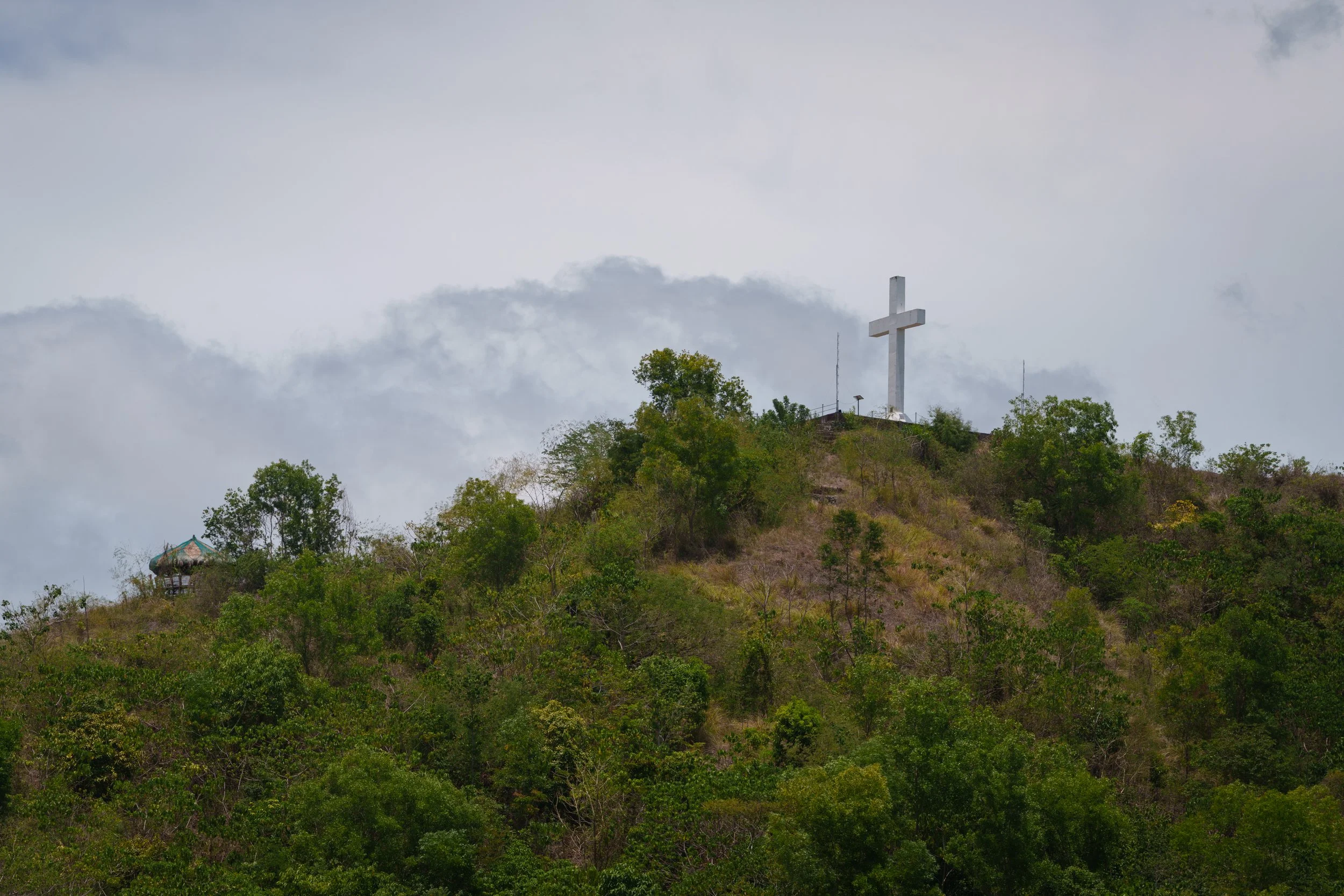 A large white cross on a hilltop surrounded by trees, with a small pagoda-style structure to the left, under a cloudy sky.