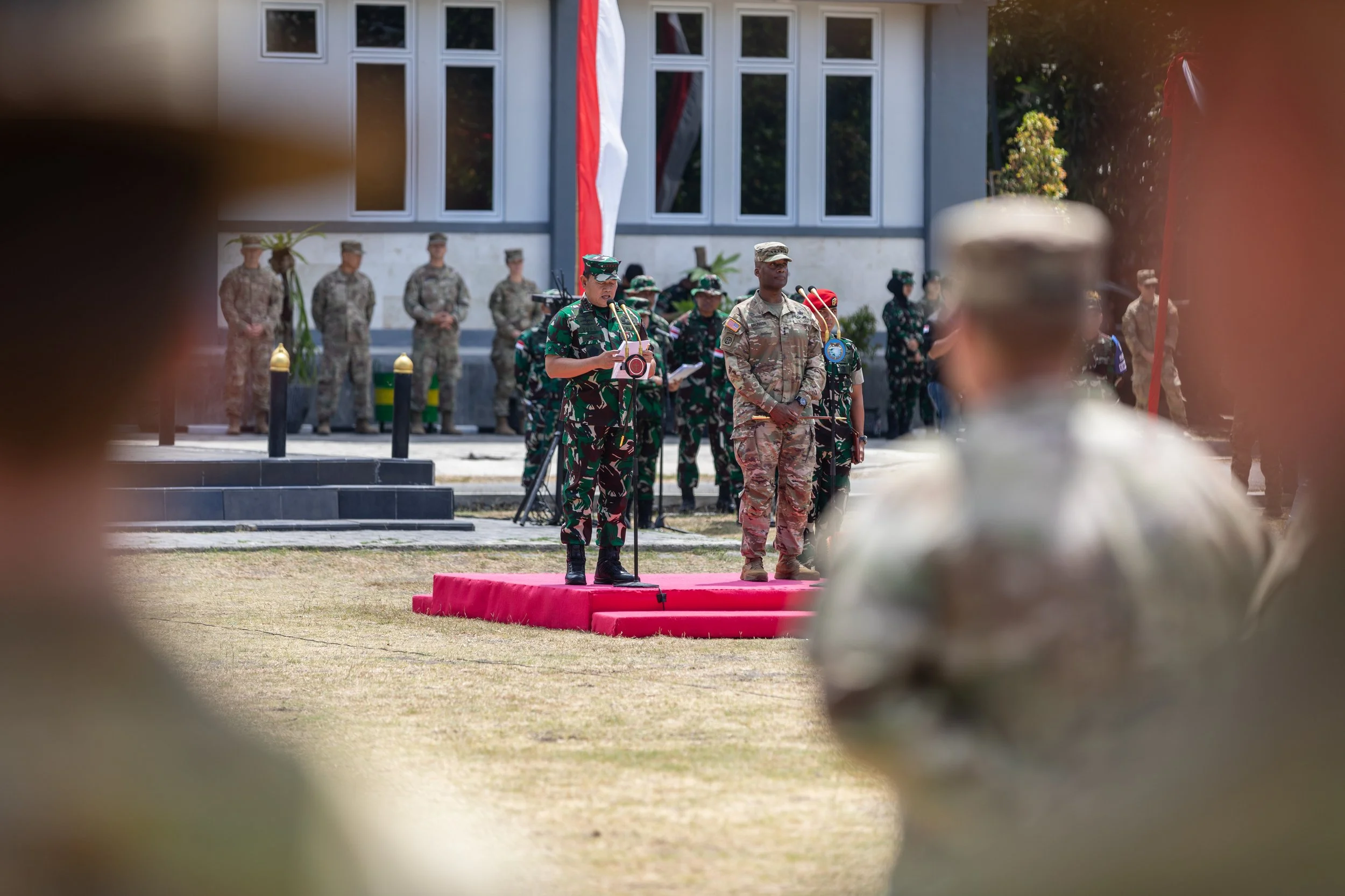 Military personnel in uniform attending a ceremony. One soldier is speaking at a podium on a pink platform, with others standing in formation behind him. The event is outdoors in front of a building with large windows, and there are black posts aroun