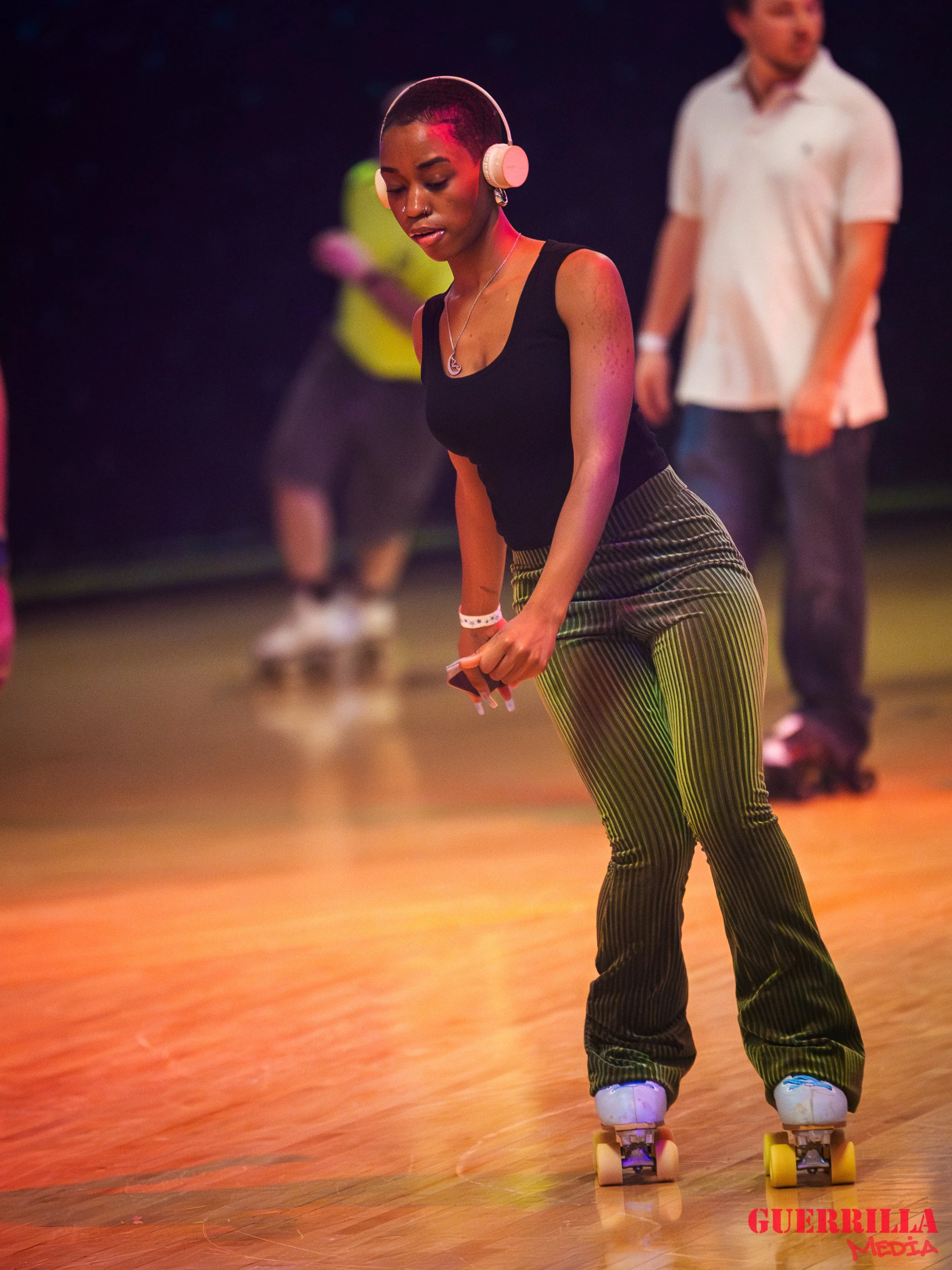 A young woman roller skating indoors with headphones on, wearing a black tank top and striped pants.