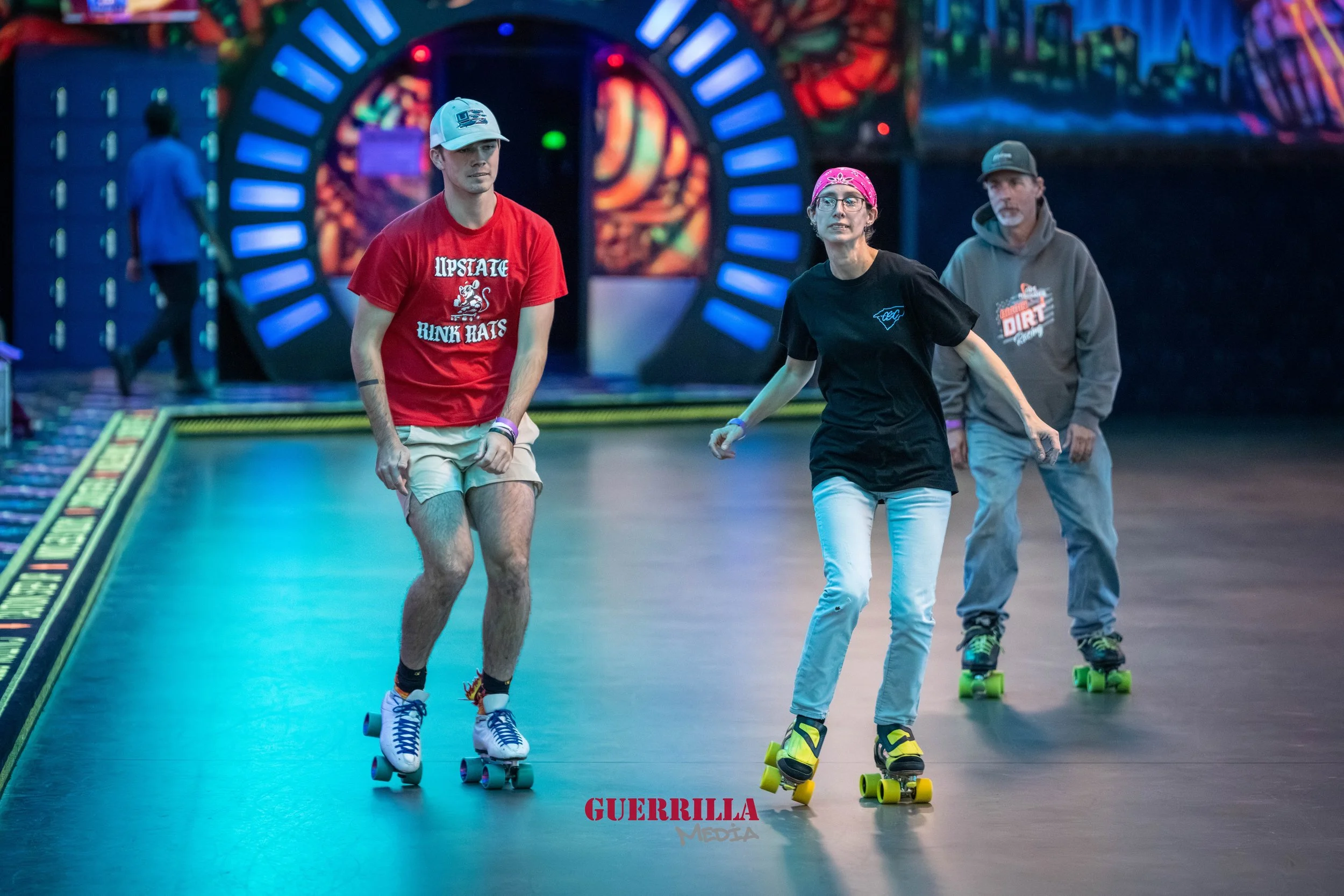 Three people roller skating inside an arcade with colorful neon lights. The woman in the middle is wearing a pink bandana and black t-shirt, the young man on the left is wearing a white cap and red t-shirt, and the man on the right is wearing a gray 